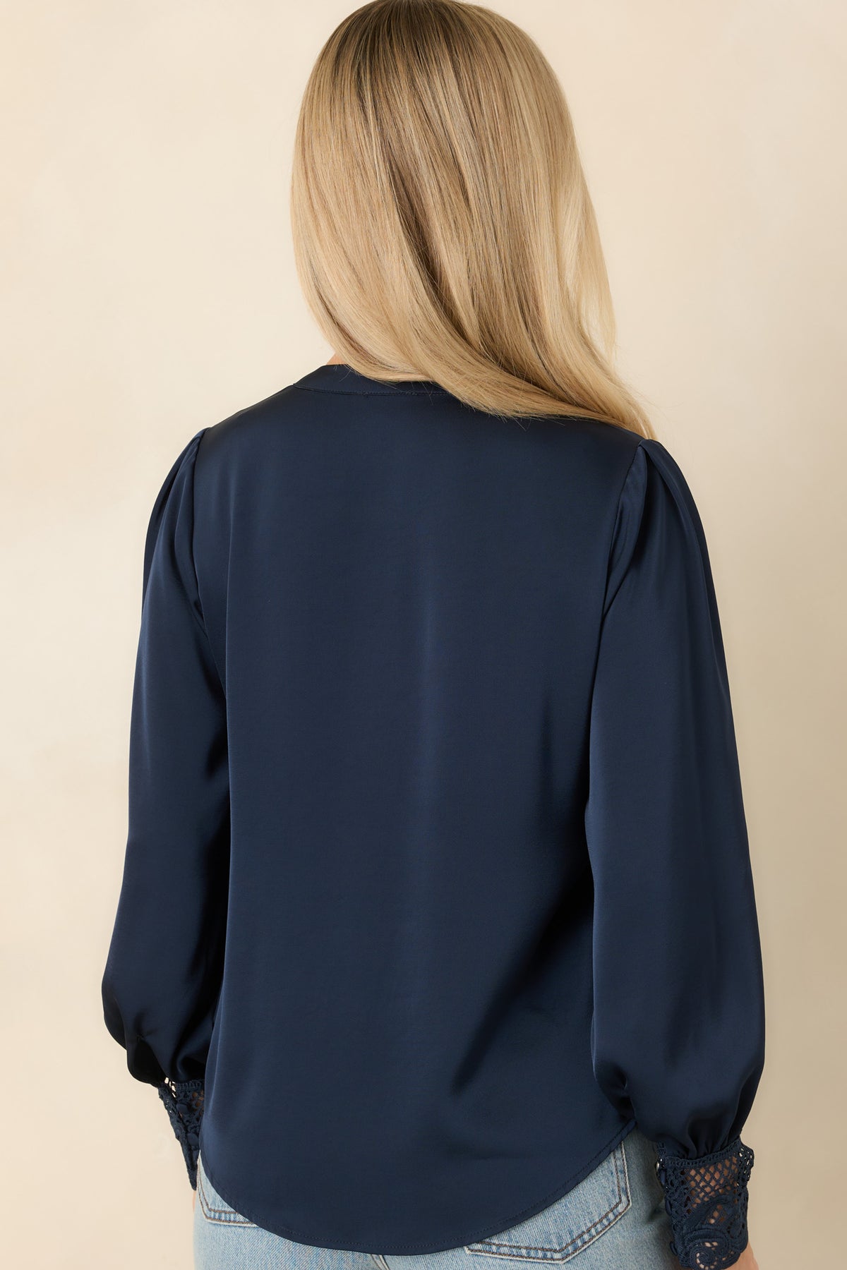 A woman viewed from the back wearing a navy blue long-sleeve blouse with sheer lace cuffs, paired with light blue jeans, against a solid beige background.