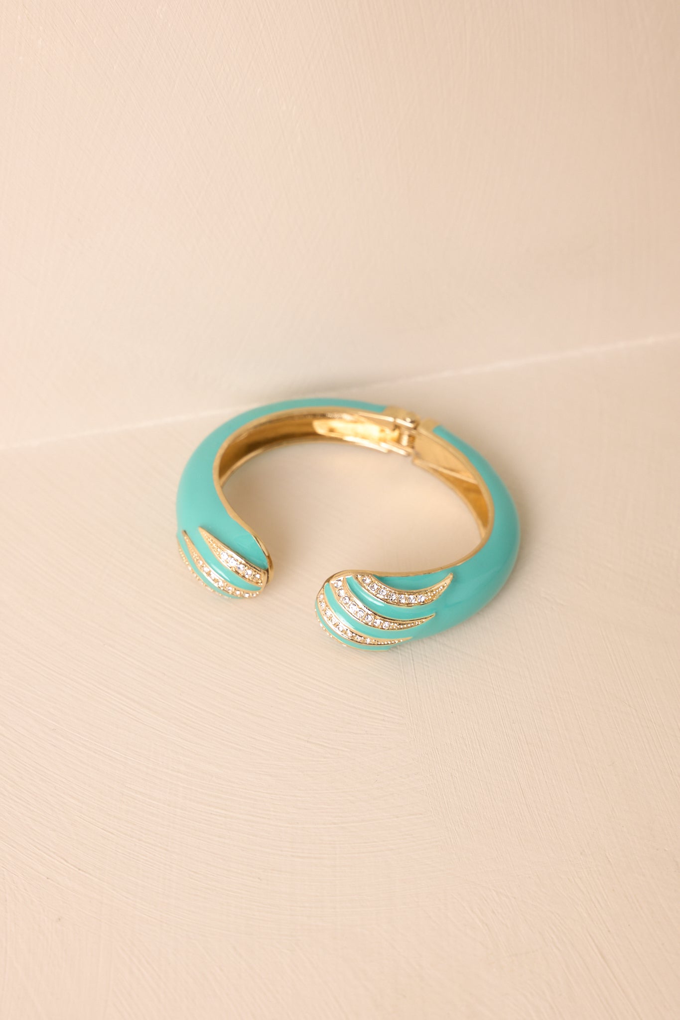 Bracelet laid on a beige surface, highlighting the glossy turquoise finish, open cuff style, and contrasting gold hardware details.