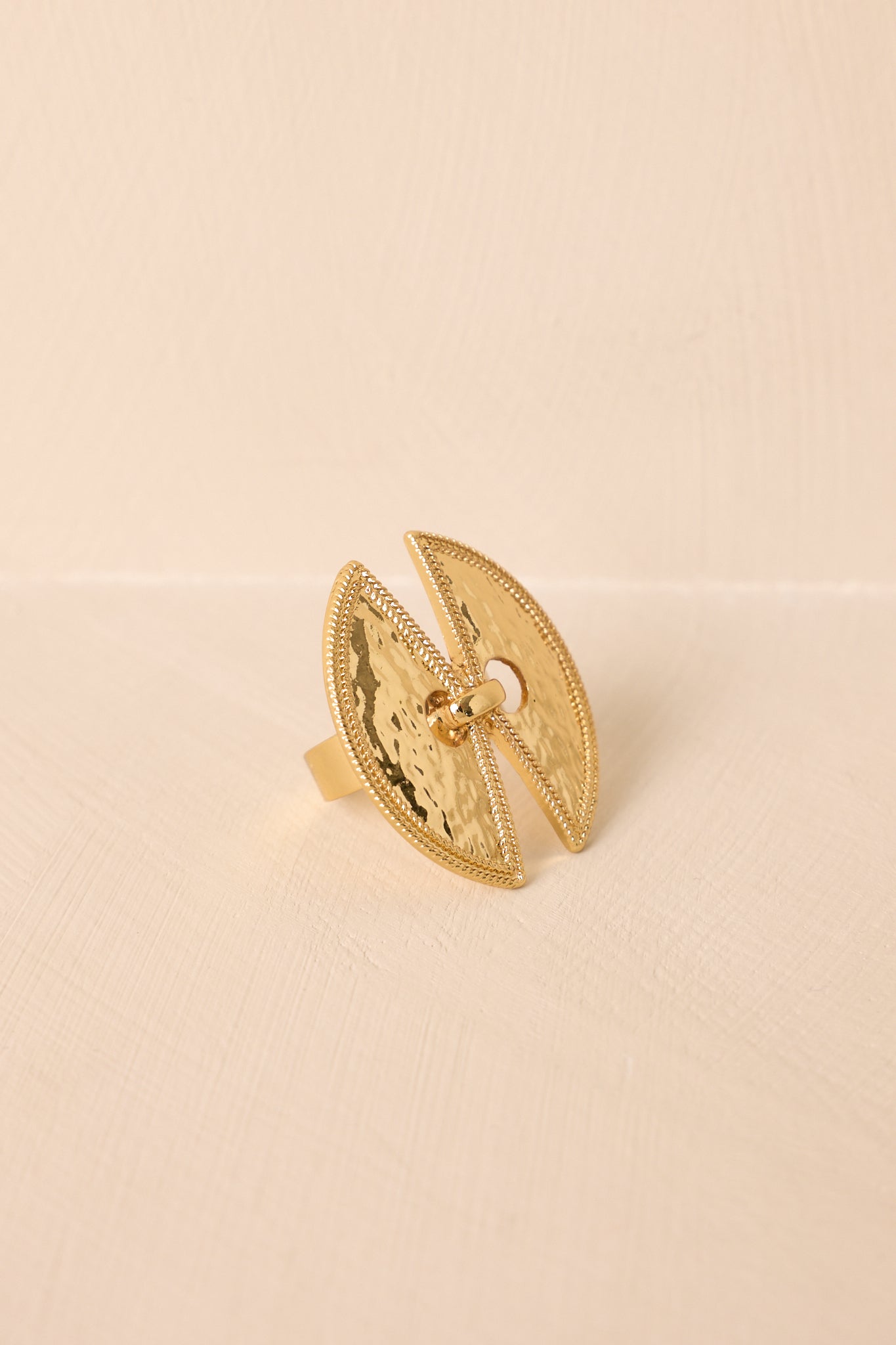 Angled shot of the ring resting on a beige surface, emphasizing the curved split design and shimmering hammered texture.