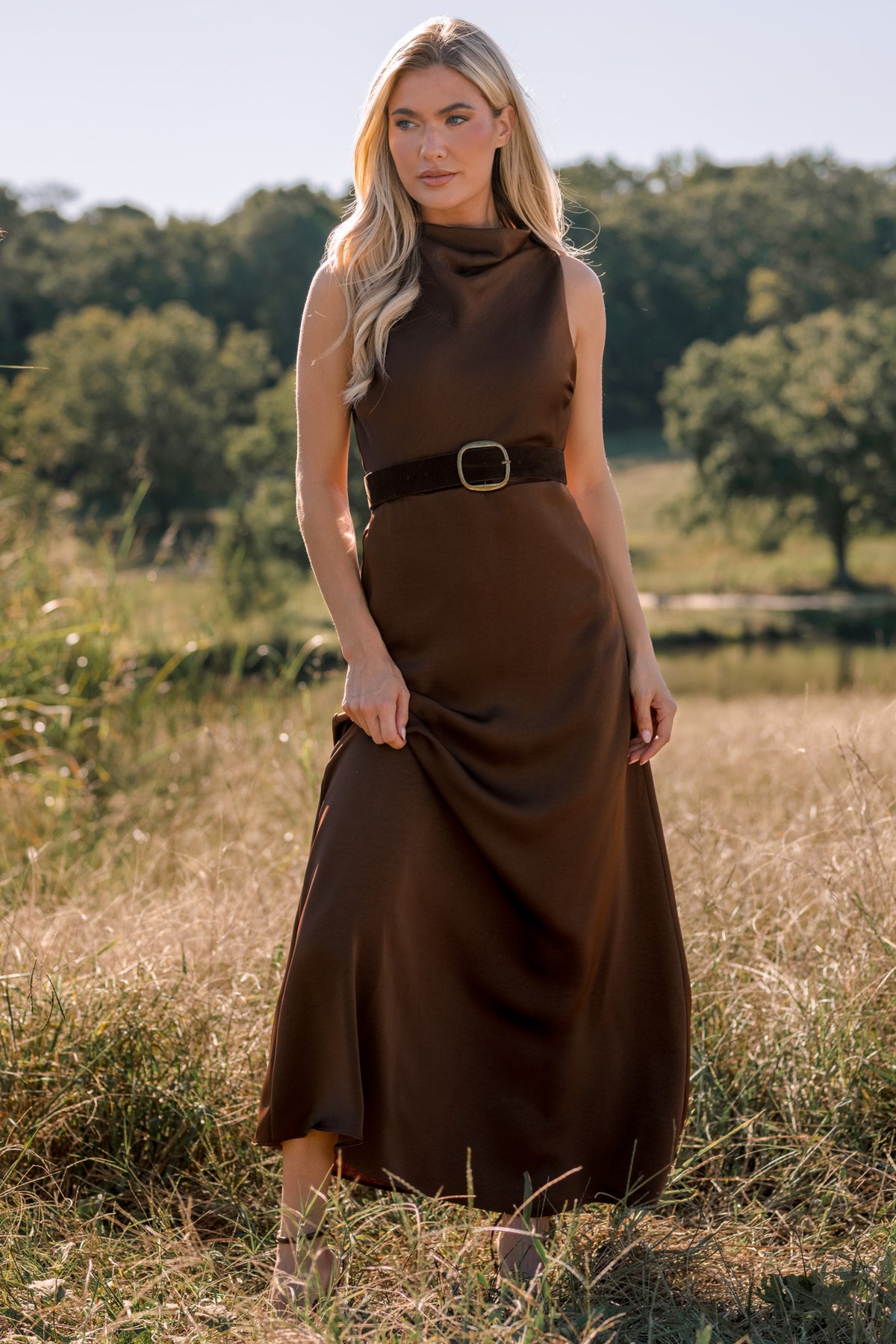 Woman modeling a sleeveless, satin brown maxi dress in a field. The dress has a belt at the waist and a cowl neckline.