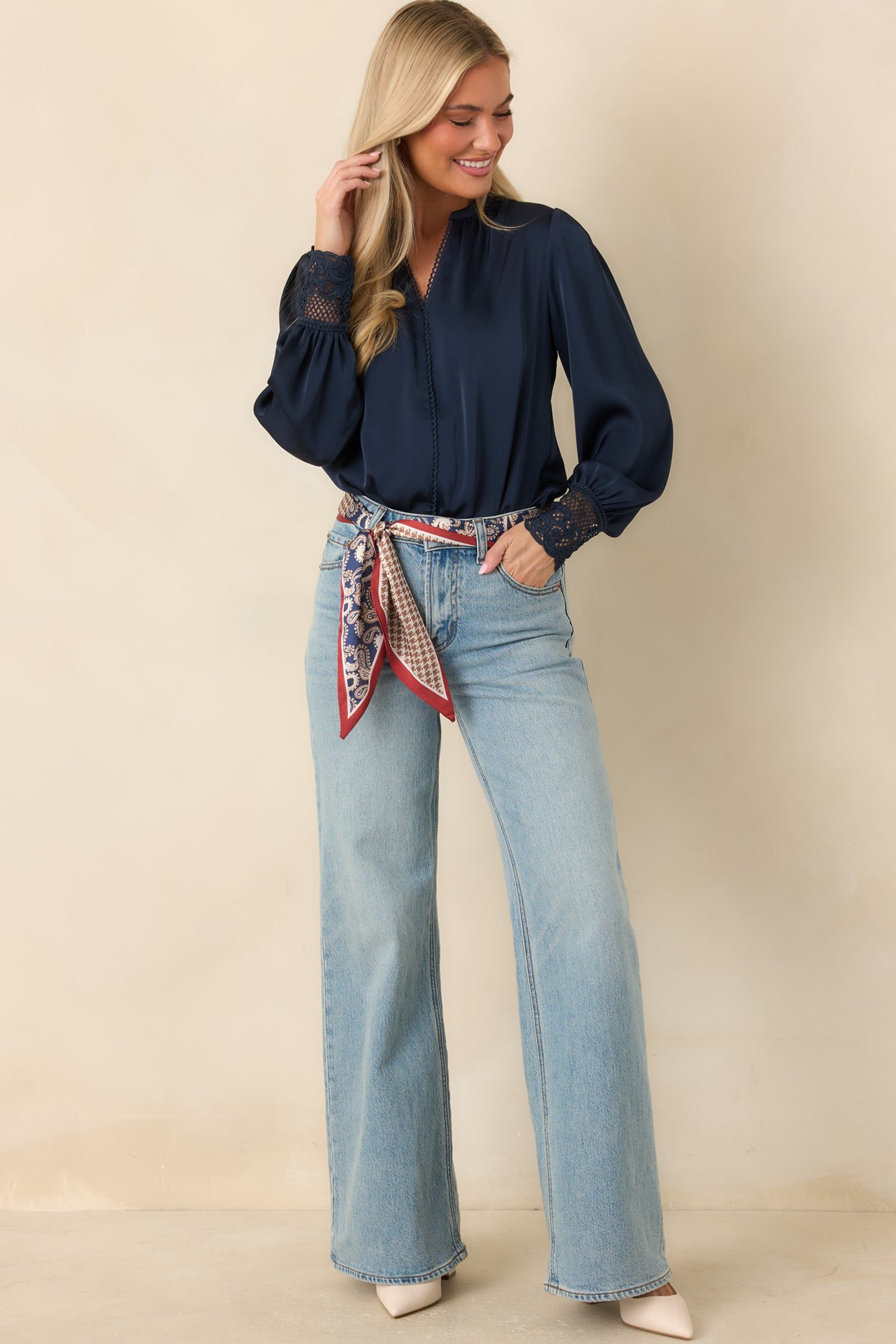 A woman modeling a navy blue long-sleeve blouse with puffed sleeves and a red, white, and blue patterned scarf tied around the waist, paired with light-wash wide-leg jeans and beige heels, against a solid beige studio background.