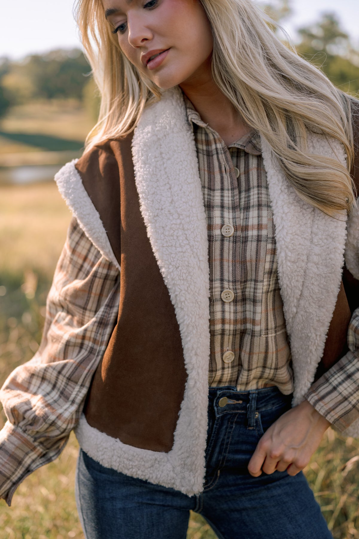 Crop in photo of a woman modeling a brown vest with ivory sherpa trim, over a beige flannel shirt, and dark skinny jeans in front of a field.