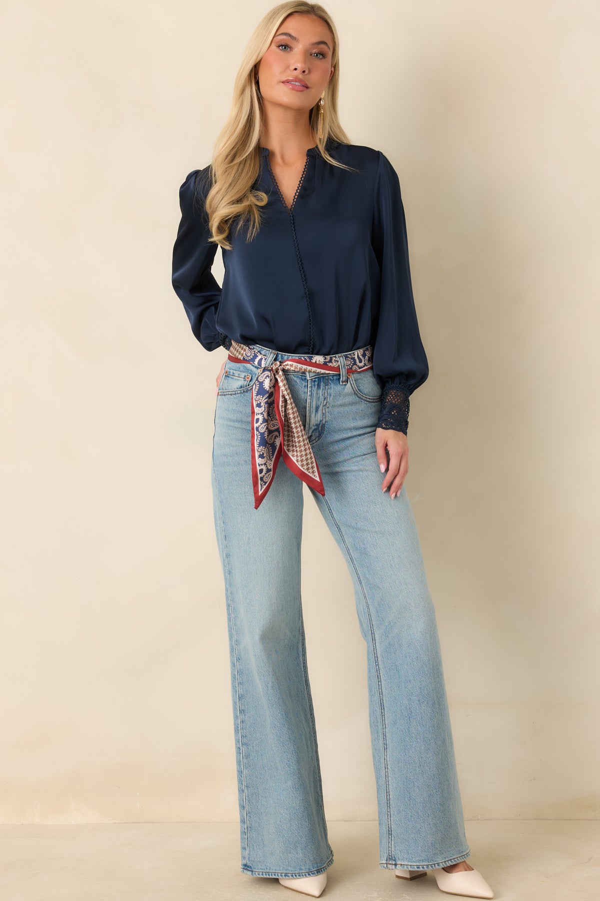 A woman modeling a navy blue long-sleeve blouse tucked into light-wash flare jeans with a red, white, and blue patterned scarf tied at the waist, against a plain beige studio background.