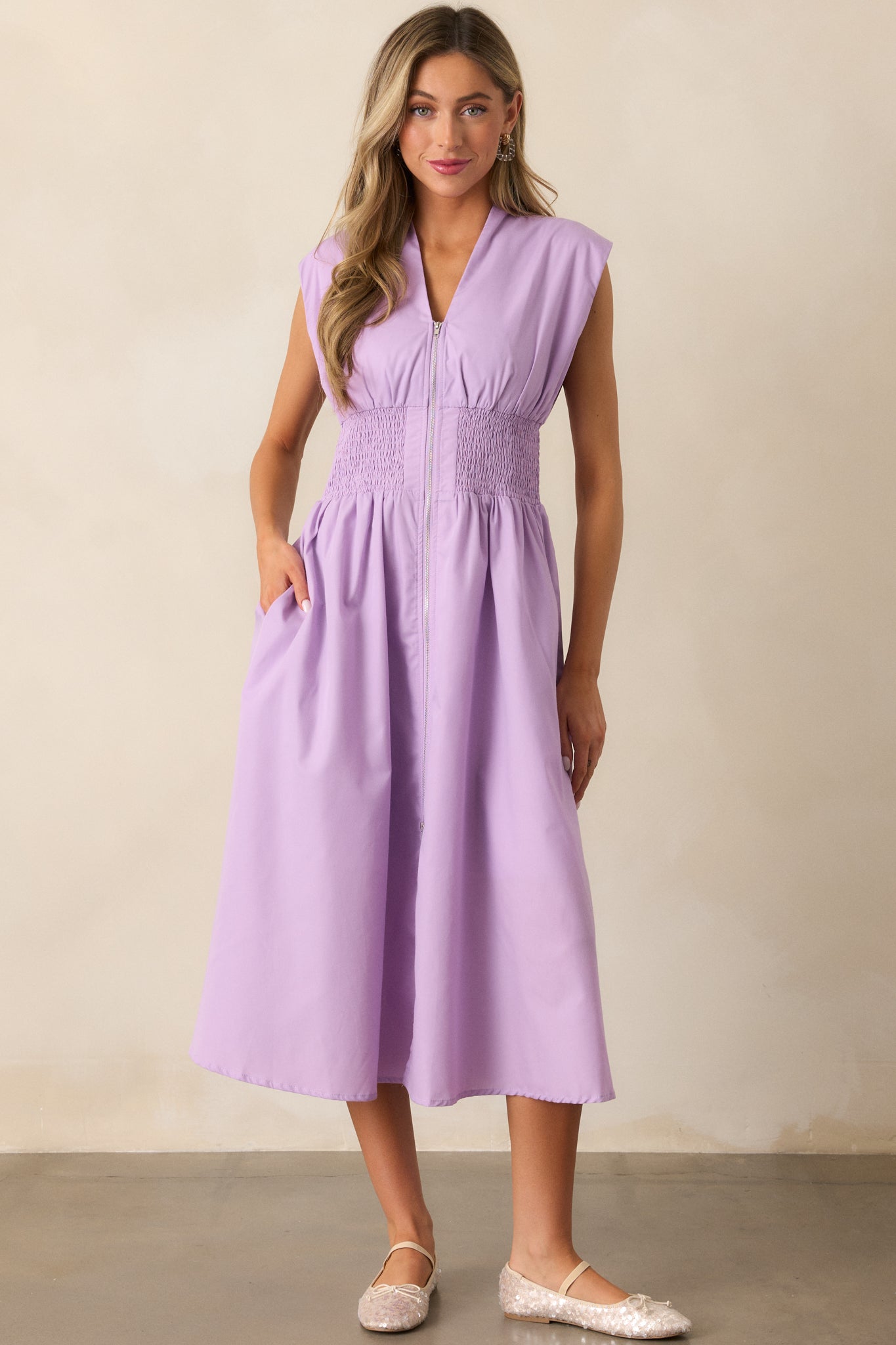 The lavender midi dress boasts padded shoulders, capped sleeves, a full zipper front, and a flattering smocked waist with functional hip pockets.