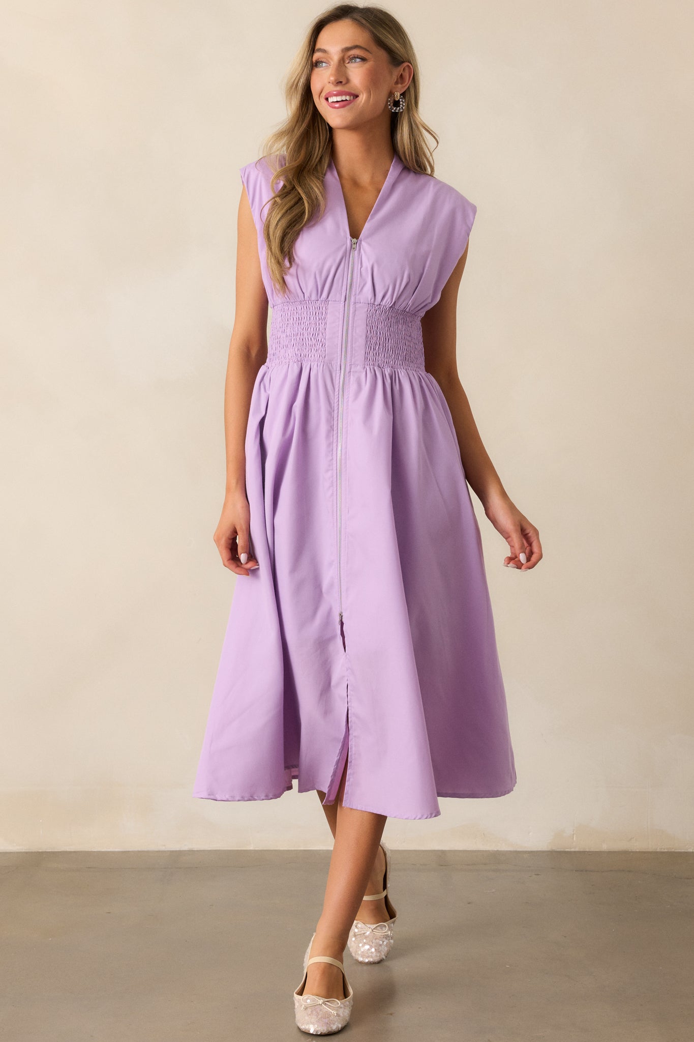 A lavender midi dress with a v-neckline, capped sleeves, and a front slit, designed with a smocked waist and functional hip pockets for a stylish, practical look.