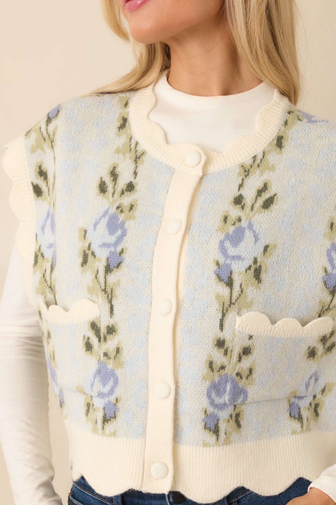 Close-up of the light blue knit vest showing scalloped trim, button-front closure, and floral textured pattern.
