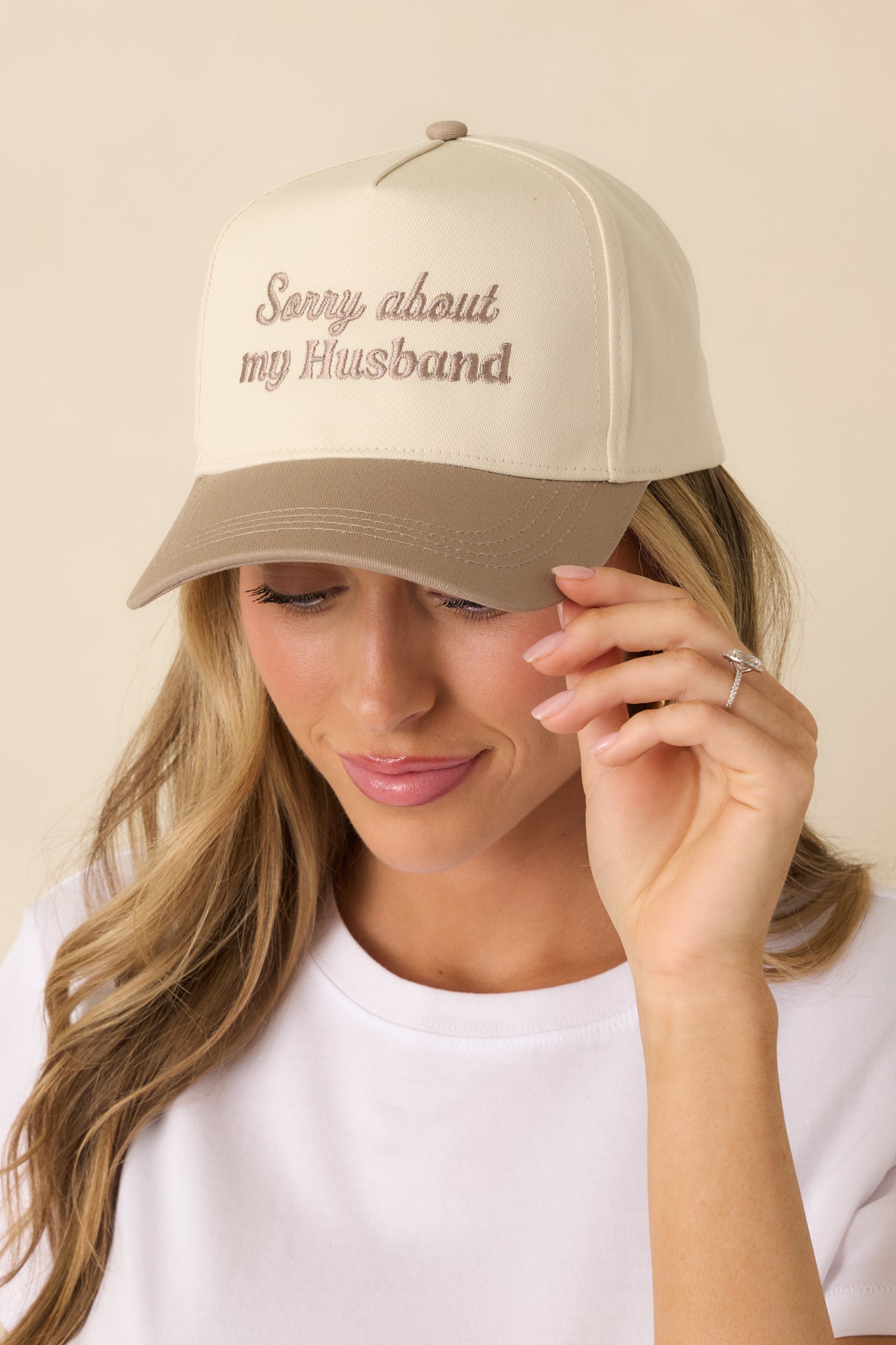Cropped view of the beige baseball cap showing the structured crown, curved brim, and tonal embroidered text on the front.