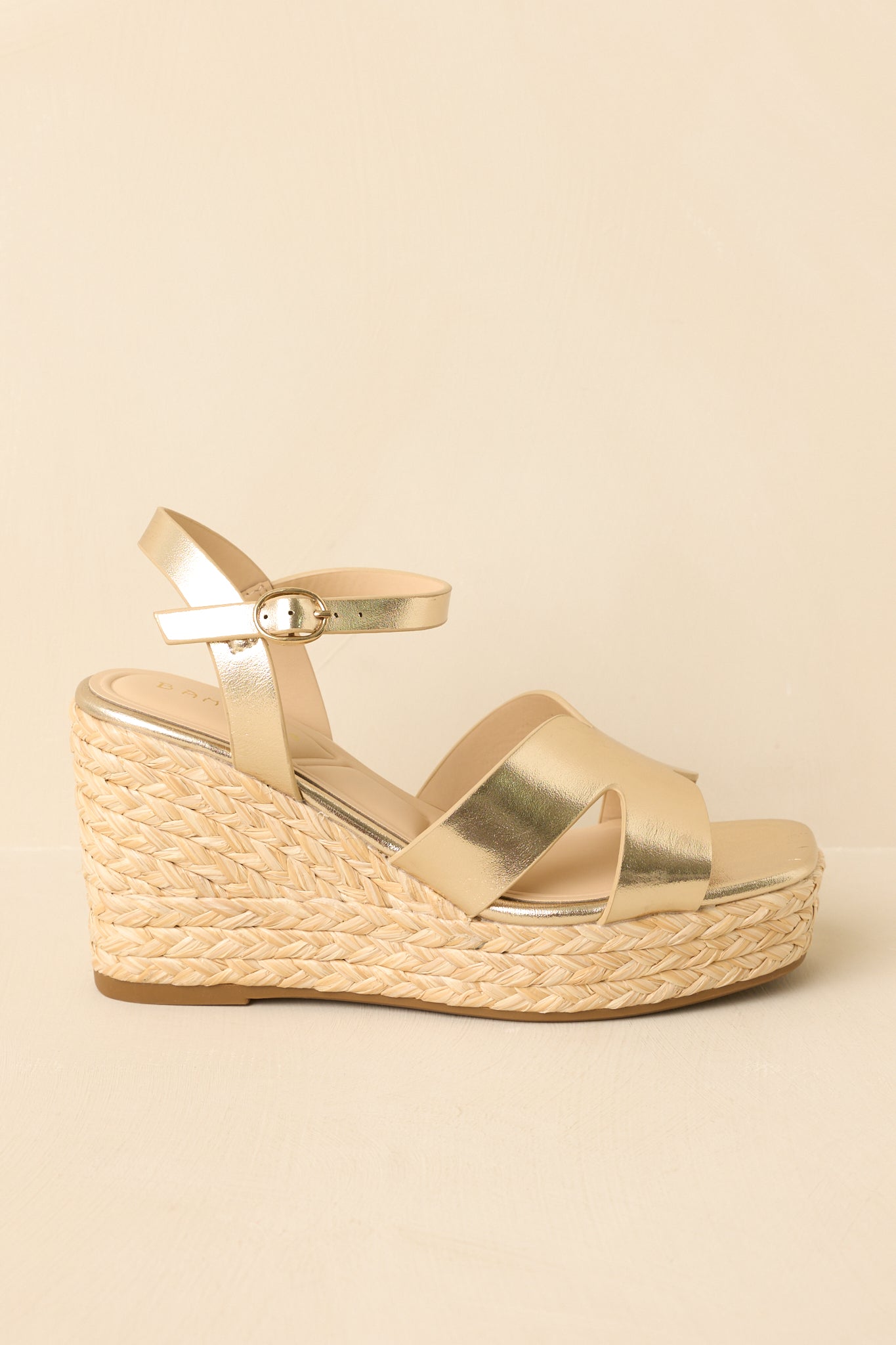 Side view of gold metallic wedge sandals on a beige background highlighting the espadrille-style platform and ankle strap.