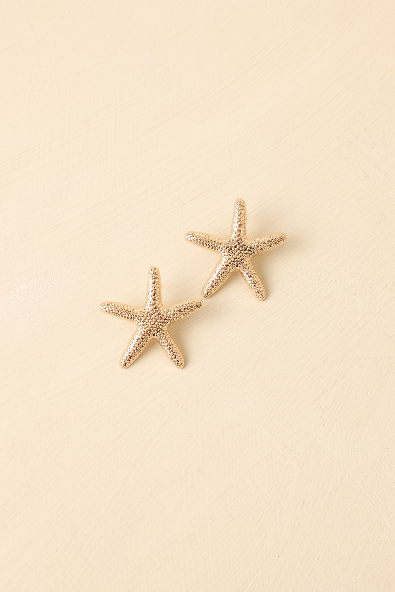 Front view of gold starfish earrings displayed on beige background emphasizing textured coastal design.