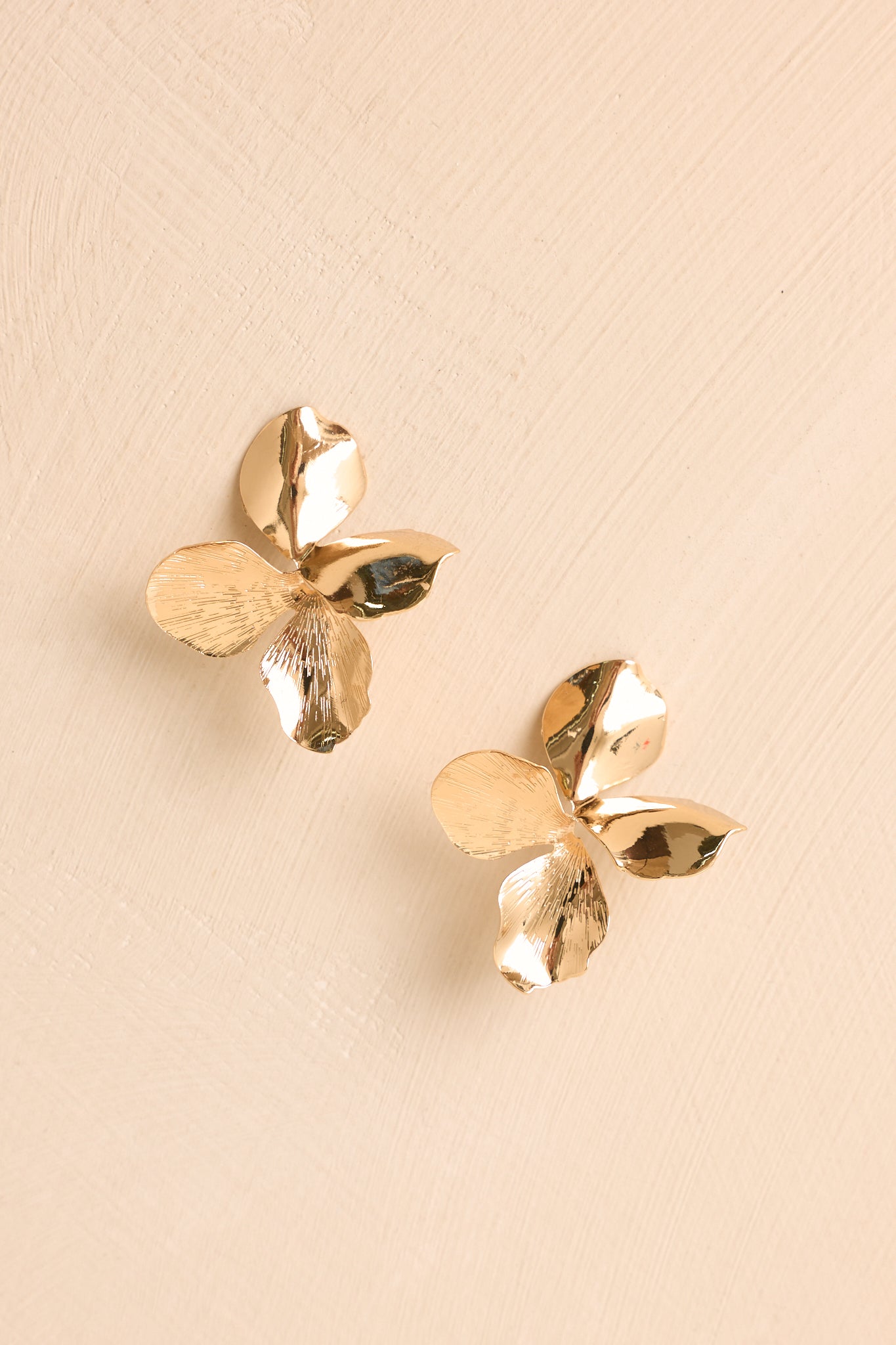 Pair of gold floral stud earrings displayed on a beige background, featuring layered petals and a polished metallic shine.