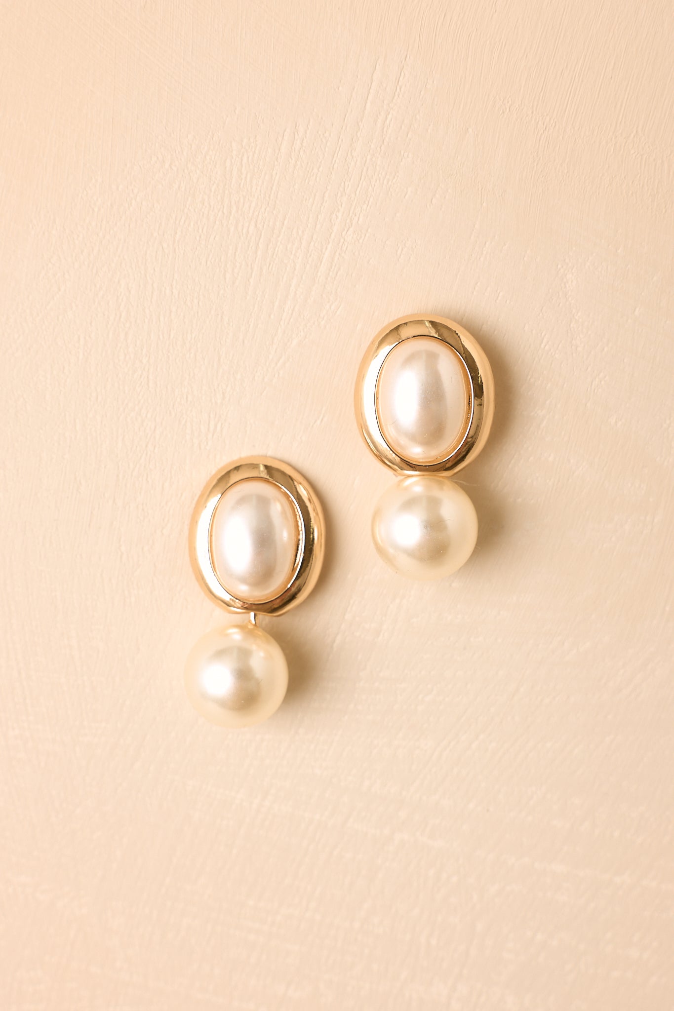 Pair of gold drop earrings displayed on a beige background, featuring oval pearl studs and coordinating pearl accents.
