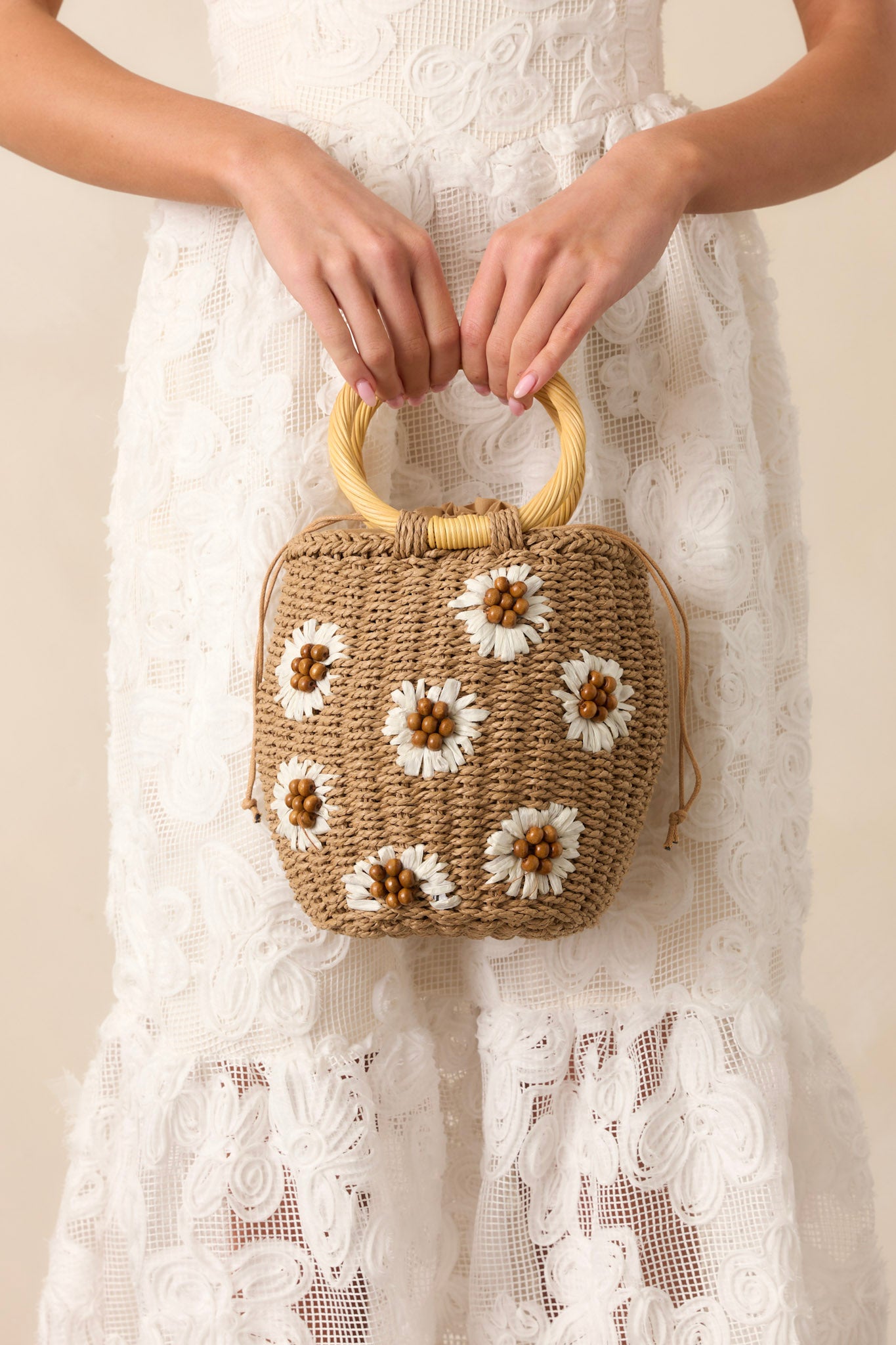 A tan straw handbag with white straw flowers and brown bead centers, featuring circular wicker handles and a drawstring closure.
