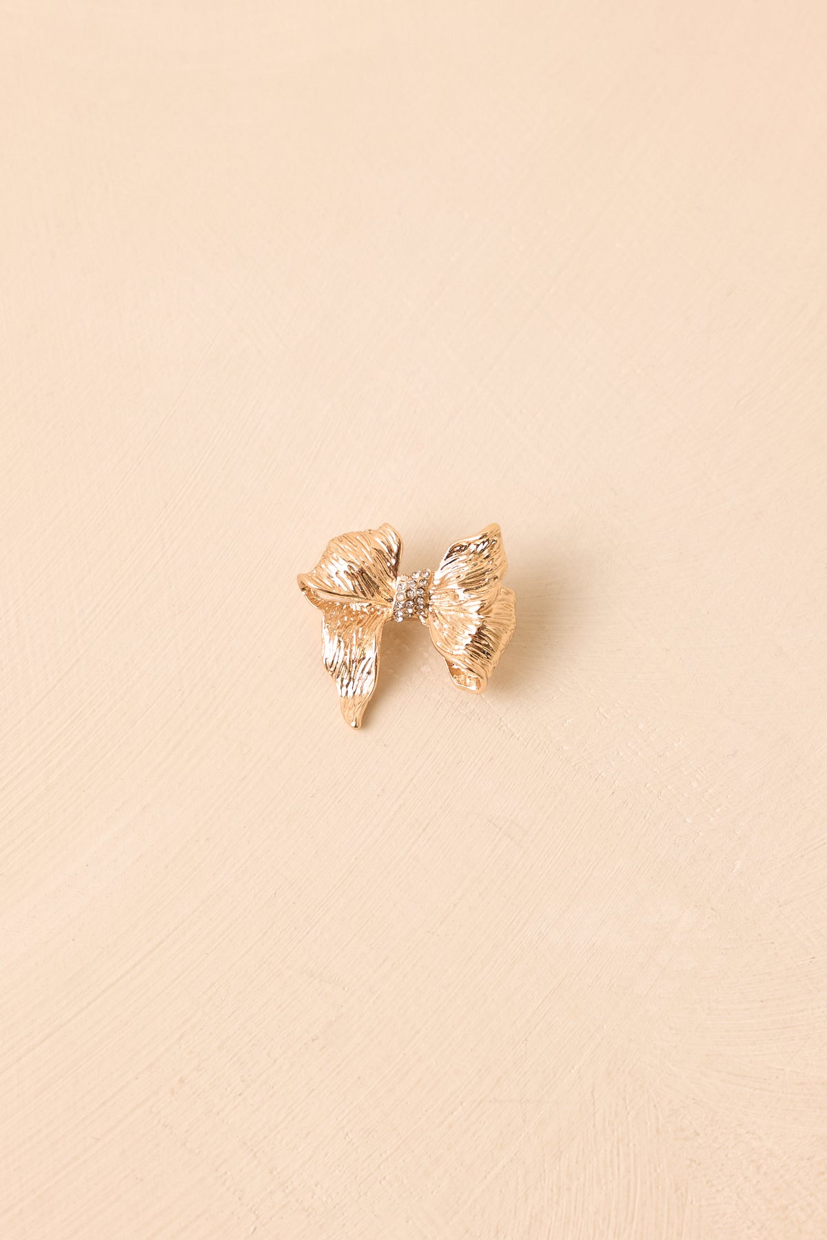 A small metallic gold bow-shaped brooch with a textured, crinkled finish, centered on a warm beige textured background.