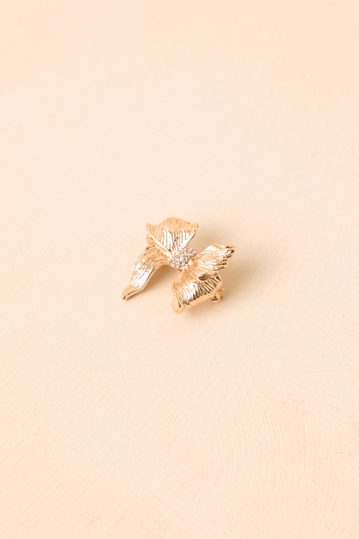 A small gold-toned brooch with a textured, leaf-like or butterfly-inspired organic shape, photographed on a plain warm beige background.