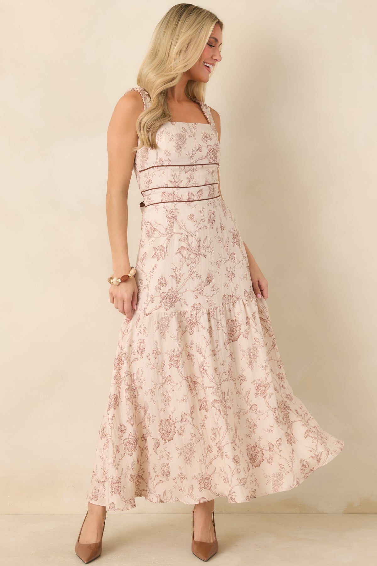 A woman modeling a sleeveless, tiered maxi dress with a dusty pink and beige floral toile pattern, standing against a plain beige studio backdrop.
