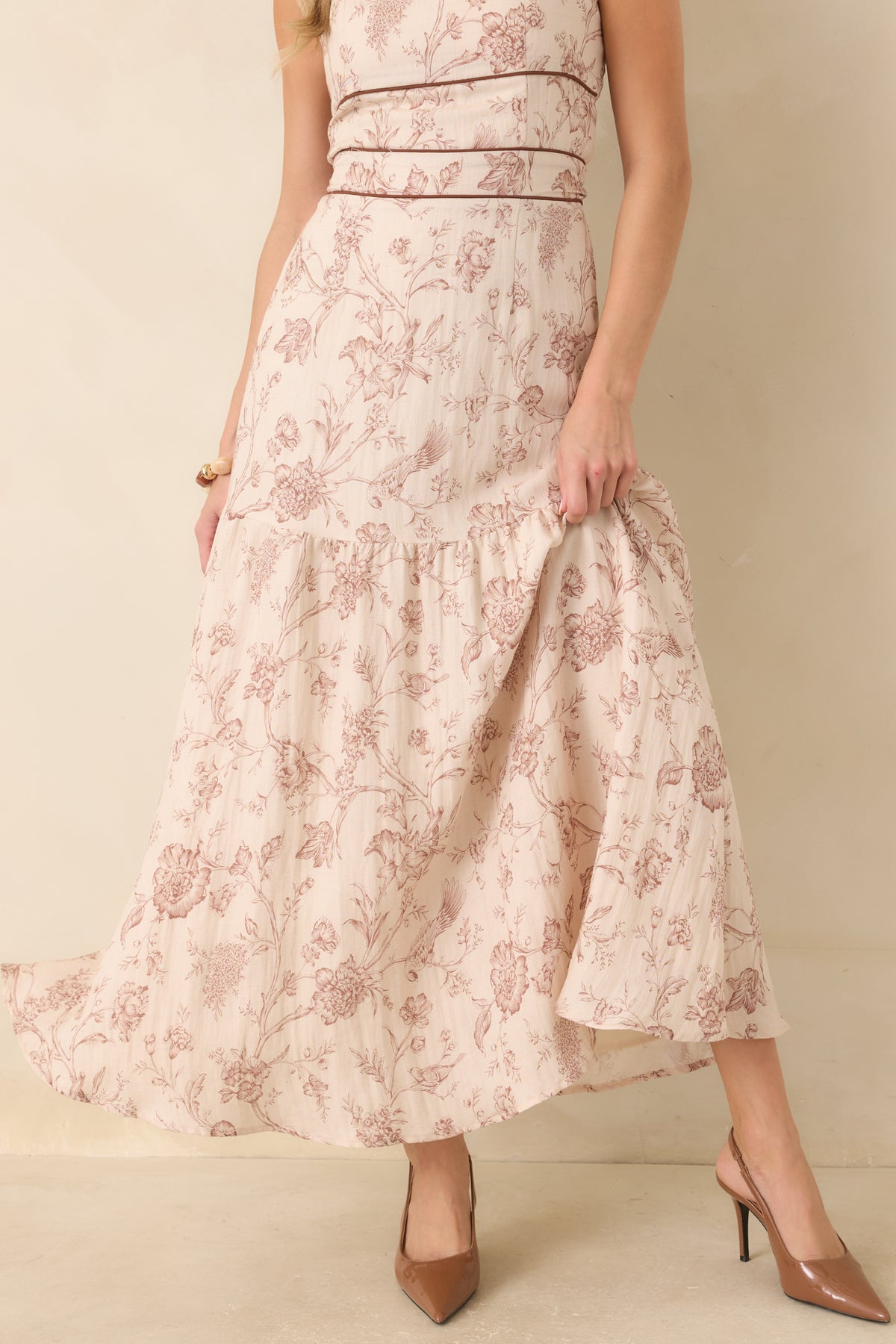 A sleeveless maxi dress with a dusty rose floral toile pattern on a cream background, shown on a model wearing brown pointed-toe heels against a neutral beige backdrop.