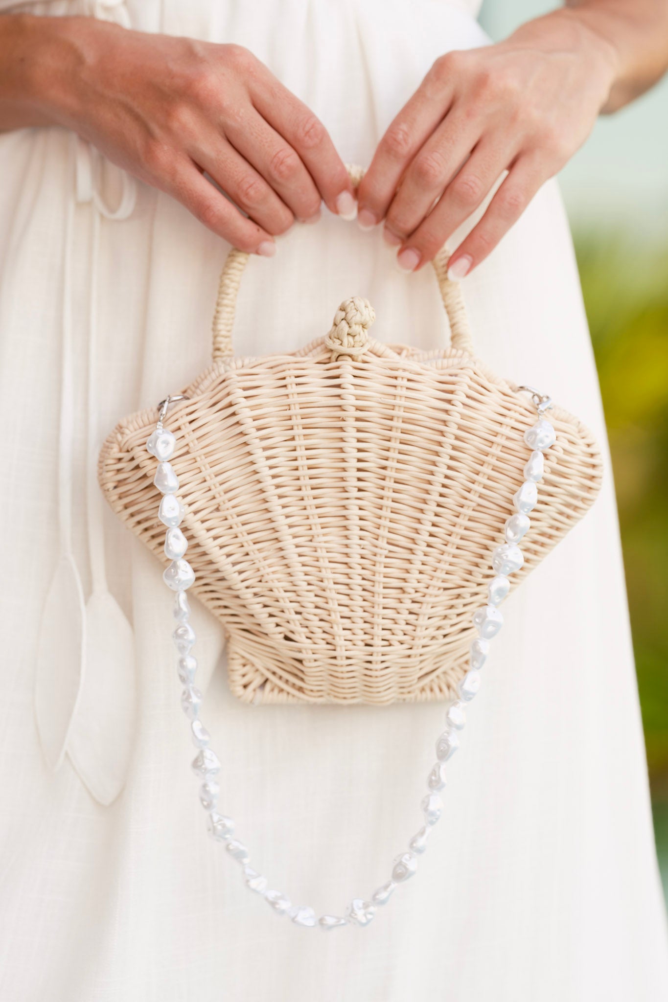 That Sunkissed Glow Ivory Rattan Structured Seashell Handbag with loop and knot toggle closure, woven handle, and ivory faux pearl shoulder strap, shown held in front of a white dress.