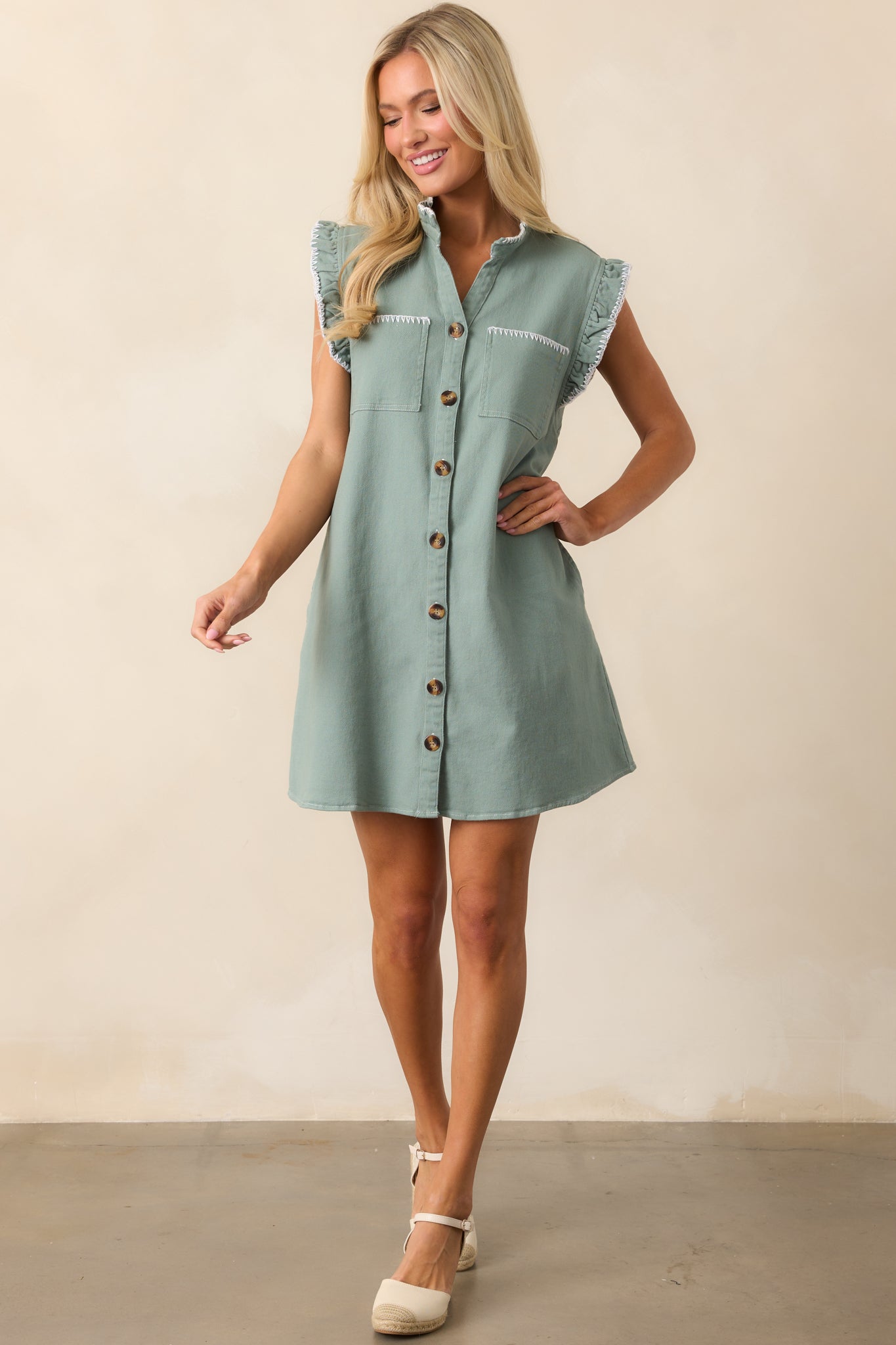 A sage green mini dress with a button front design, functional pockets, and ruffle accents at the neckline and sleeves, completed with white stitching along the hem for extra detail.