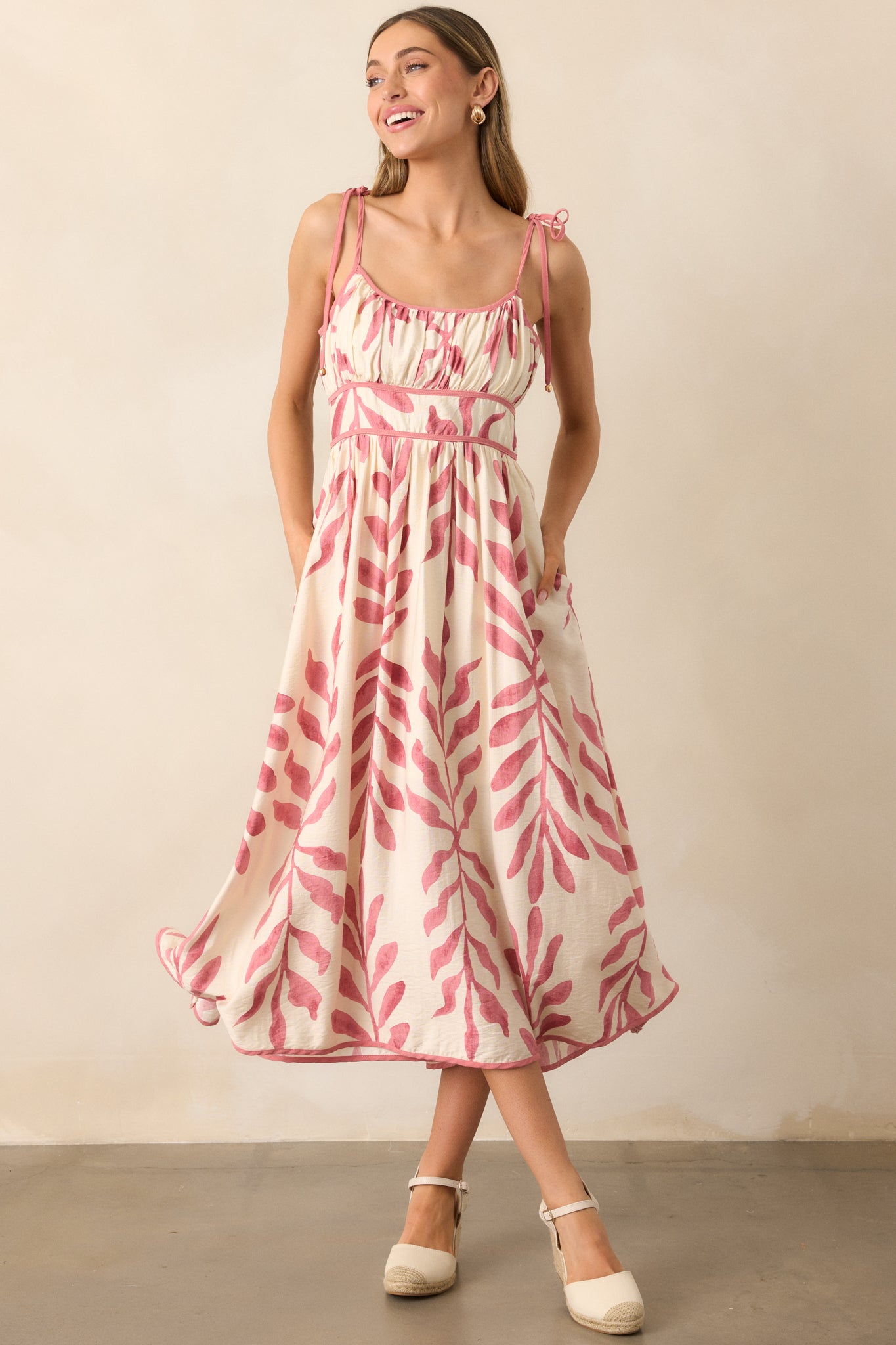 A pink midi dress with a rounded neckline, tropical palm print, and delicate self-tie spaghetti straps with gold detailing.