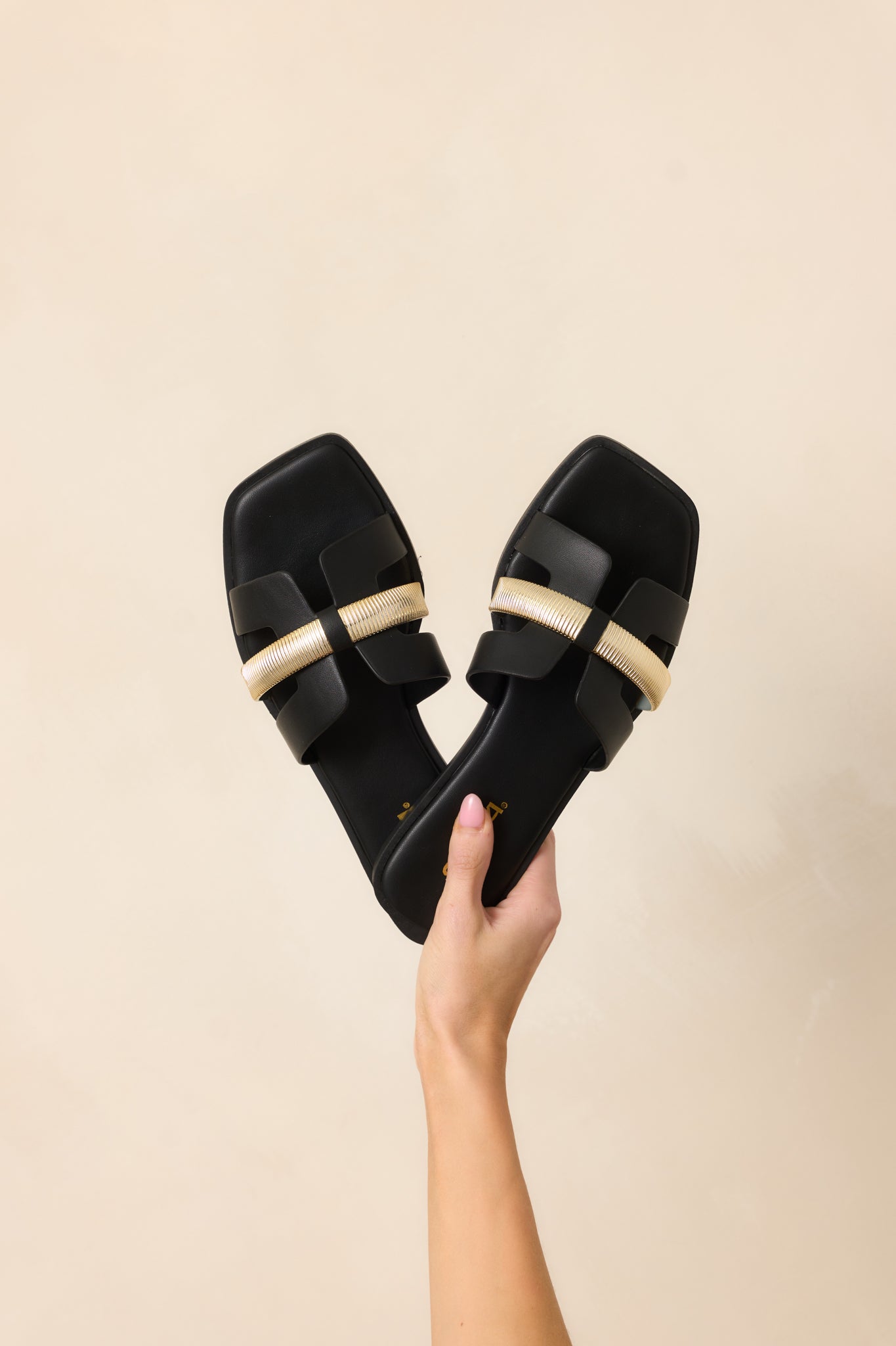 Close-up view of the black slide-on sandals, highlighting the gold accent across the vamp and the sleek faux leather material.