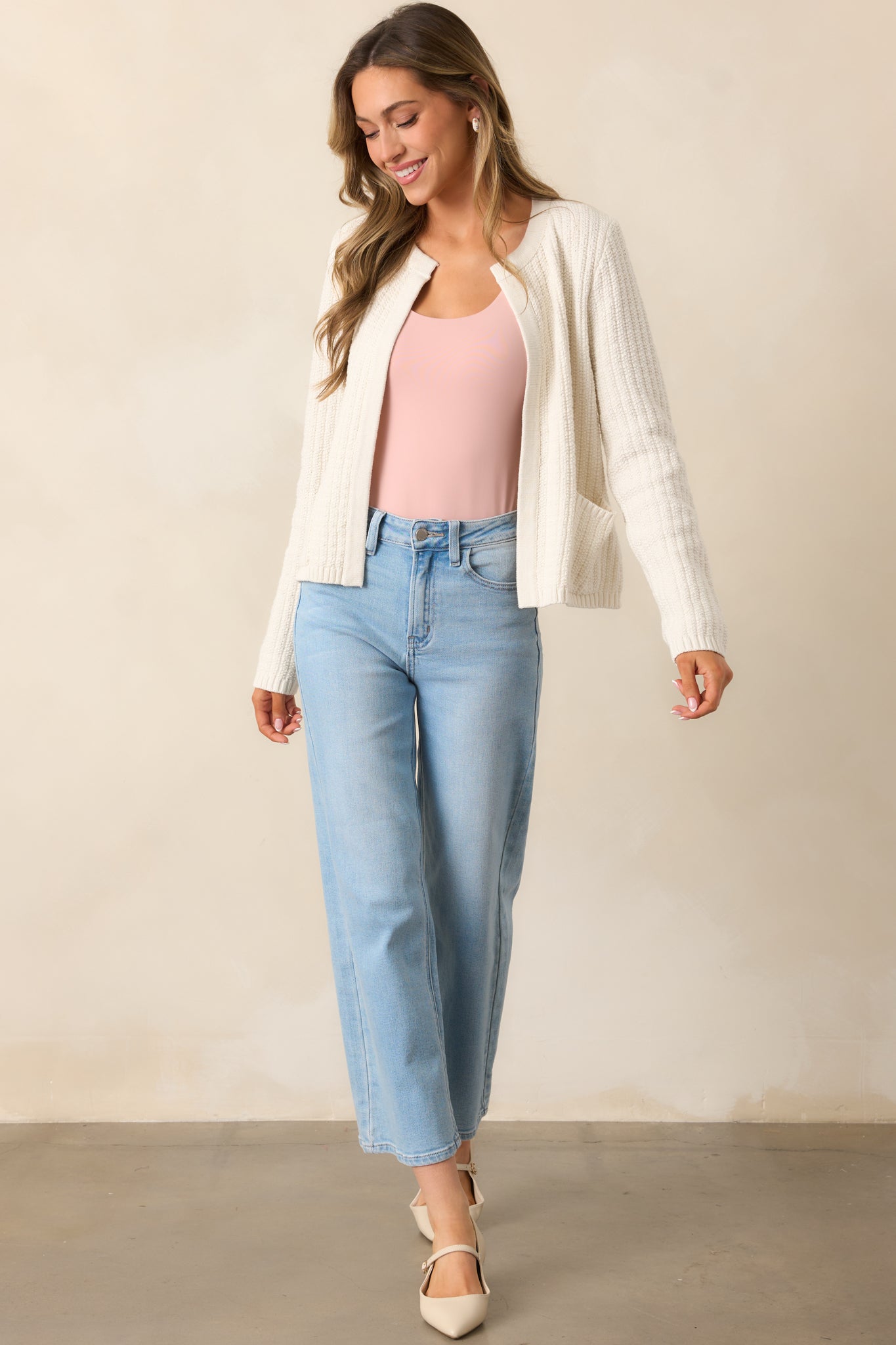 Full-length image of an ivory cardigan featuring a knit texture, long sleeves, and a rounded neckline, styled with the pockets slightly in use.