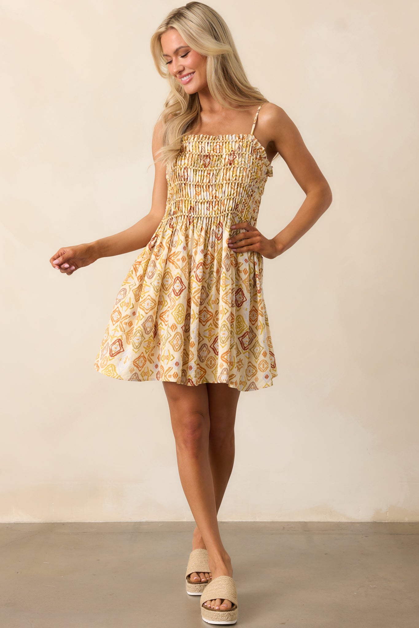 A beige mini dress featuring a square neckline, thin adjustable straps, a smocked pleated bodice with beaded detailing, and a flowing skirt.
