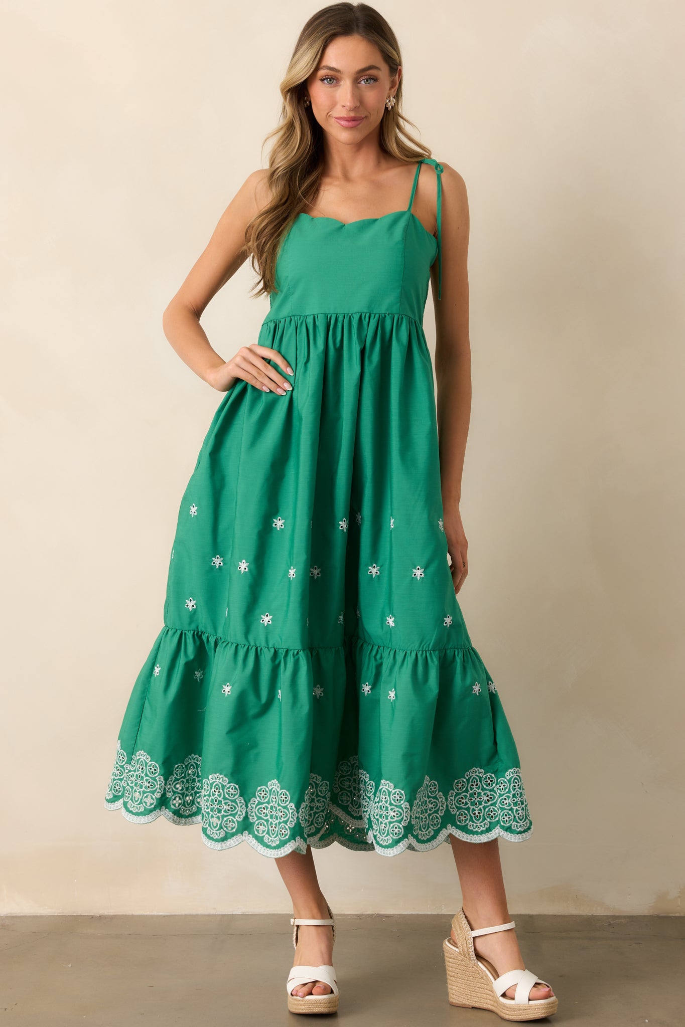 A green midi dress with a scalloped sweetheart neckline, adjustable self-tie bow straps, and a tiered skirt, featuring delicate embroidered eyelet detailing throughout.