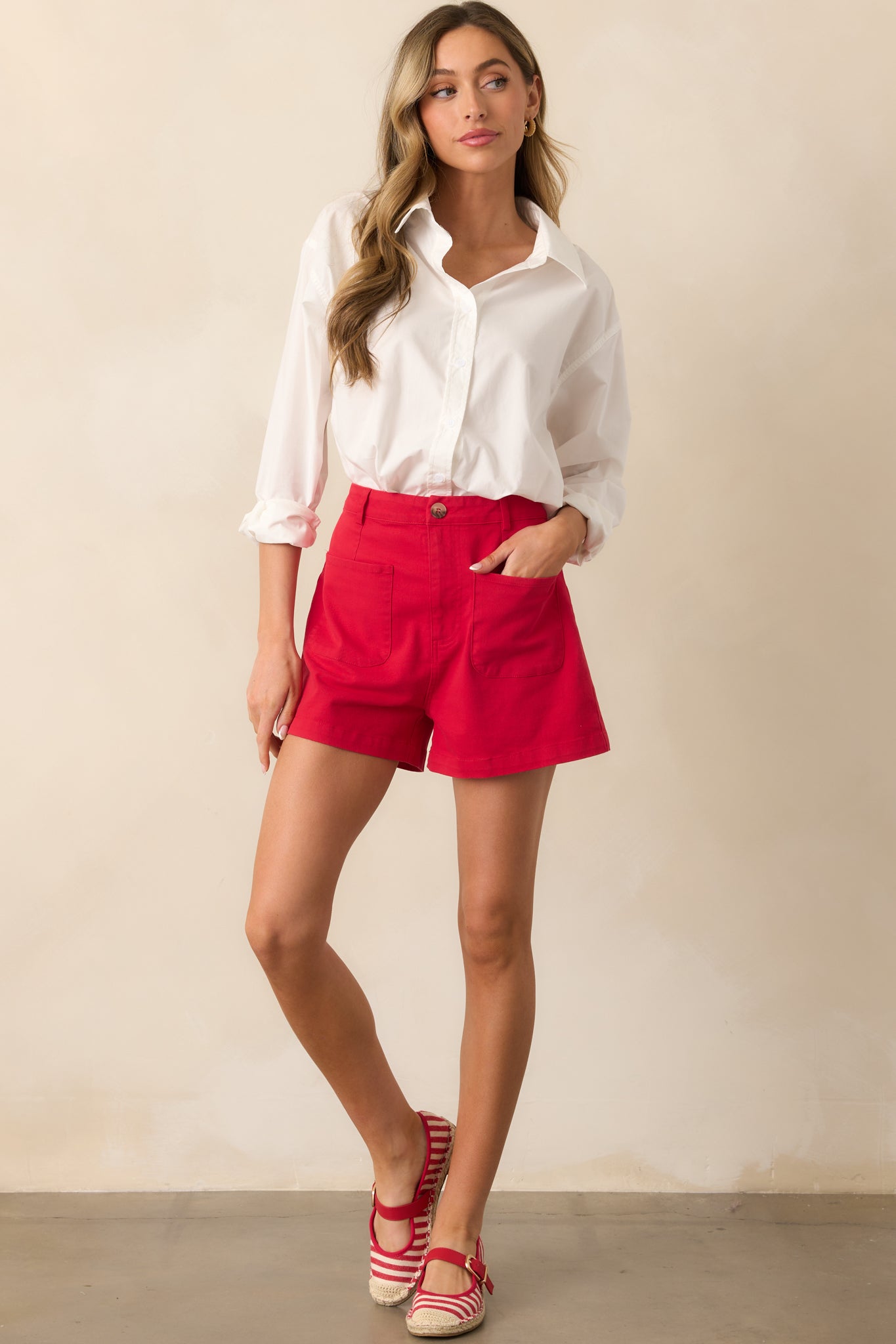 A full view of the red shorts featuring a high-waisted design, zipper and button closure with a tortoise shell button, and functional front patch pockets for a bold yet classic look.