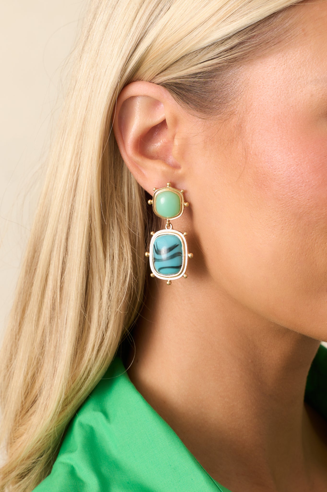 A pair of gold and turquoise drop earrings featuring gold hardware, an oval turquoise green stud surrounded by gold beading, with a rectangular sky blue marbled stone connected by gold jump rings.
