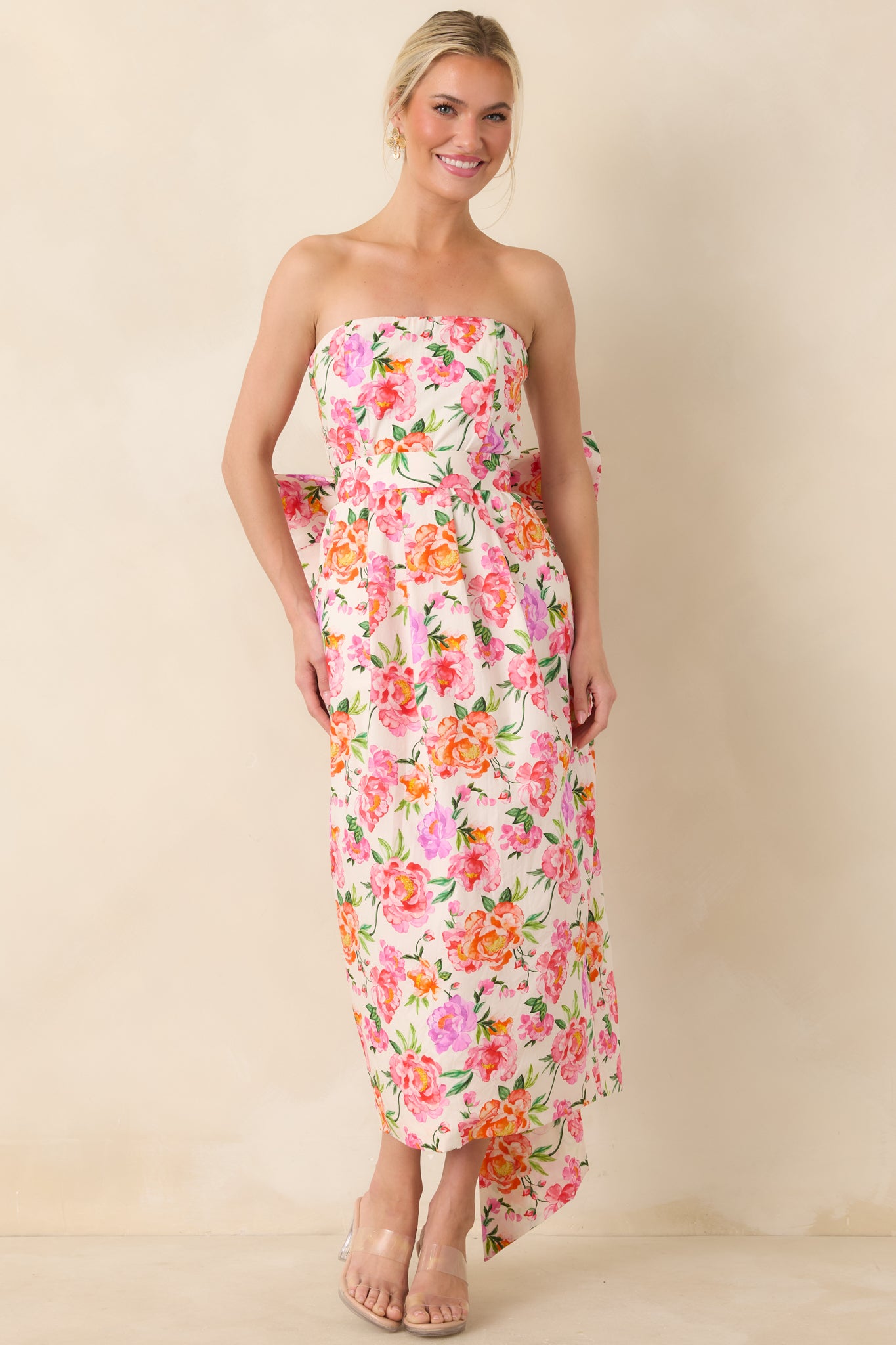 Pink floral midi dress featuring a strapless neckline, fitted bodice, and flowing skirt adorned with a playful oversized bow at the back.