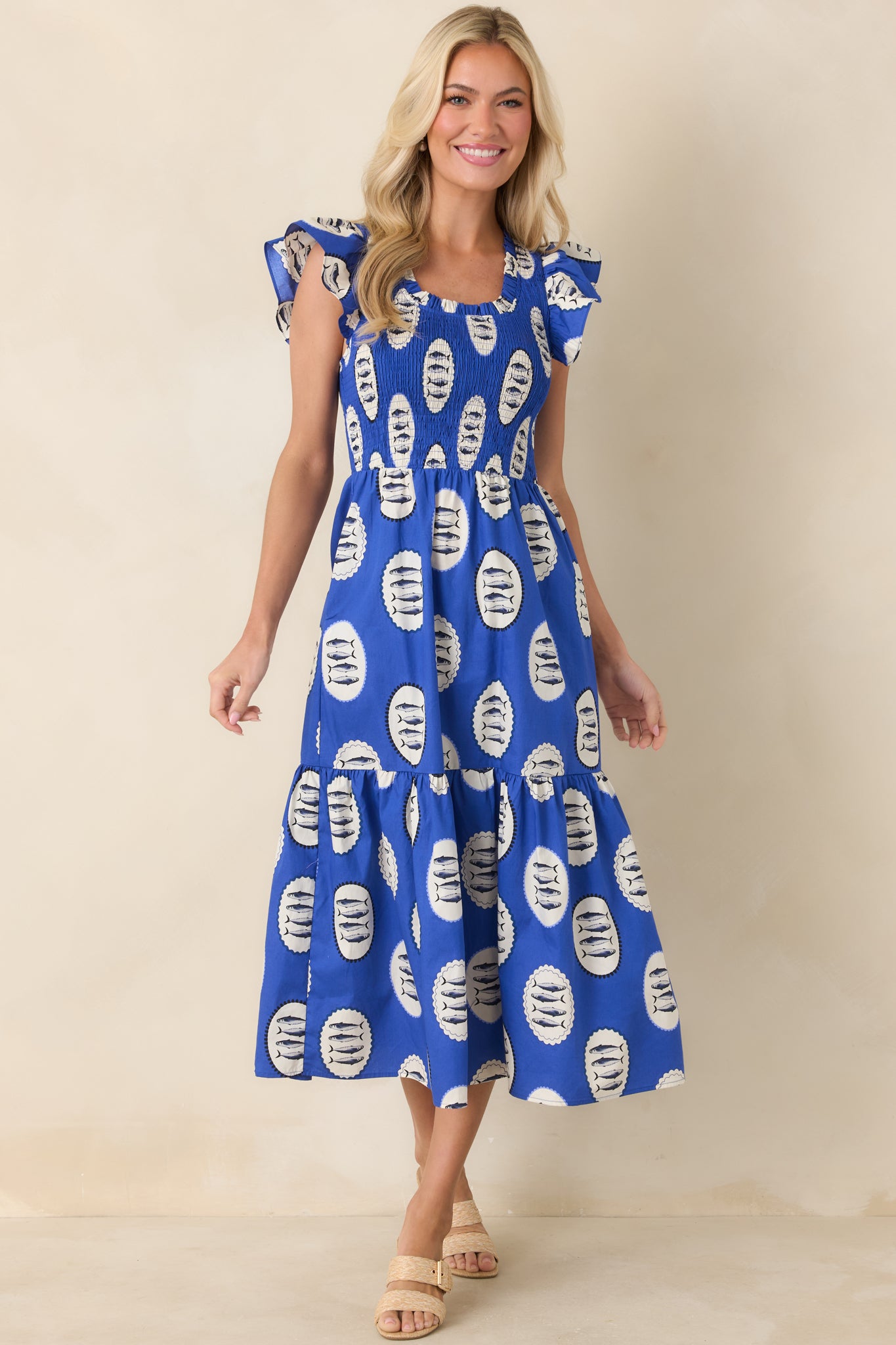 Vibrant blue cotton midi dress covered in whimsical fish motifs, designed with a stretchy smocked bodice and airy flutter sleeves above a relaxed tiered silhouette.