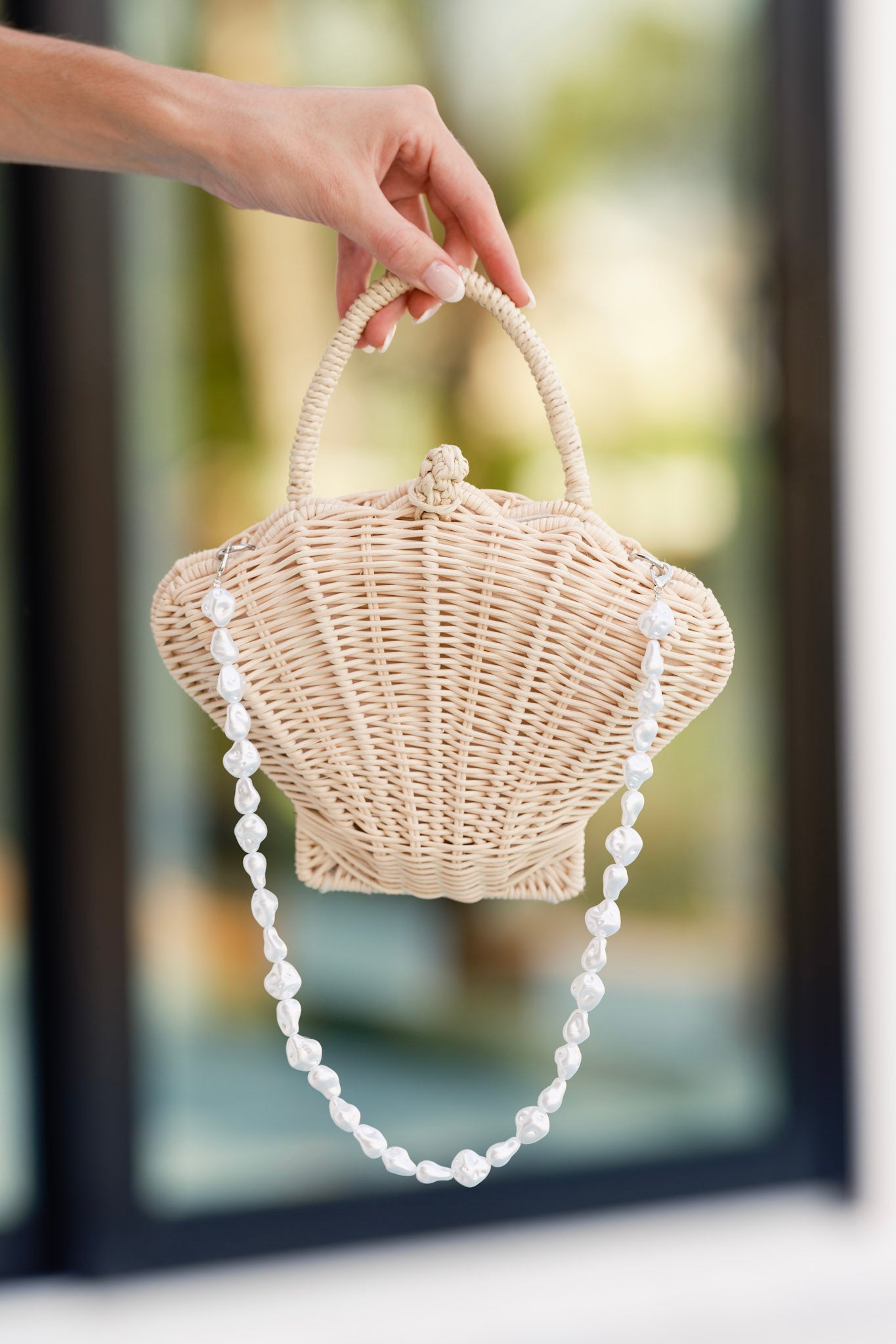 That Sunkissed Glow Ivory Rattan Structured Seashell Handbag featuring a loop and knot toggle closure, woven handle, and ivory faux pearl strap, displayed against an outdoor background.