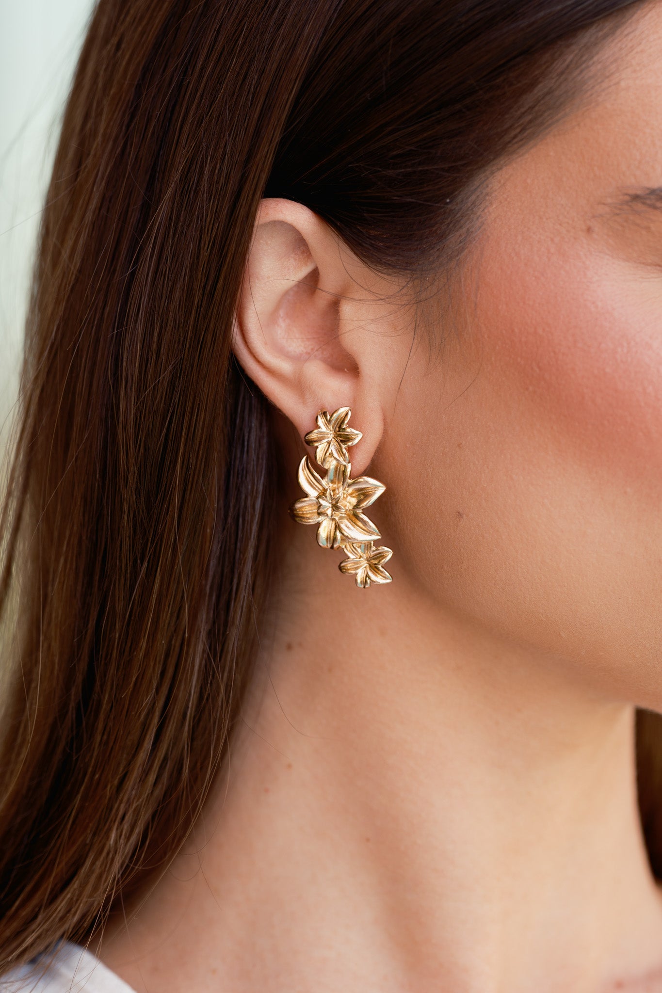 Gold earrings with a textured finish, featuring three cascading cherry blossom flowers in a drop design, shown worn on a model’s ear.