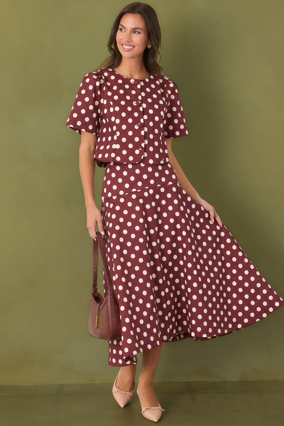 Woman modeling a two piece wine red set with ivory polka dots. The blouse is short sleeved, structured, and has buttons up the front. The bottom half is a midi skirt that is fitted at the waist and flows out, starting at the hips.