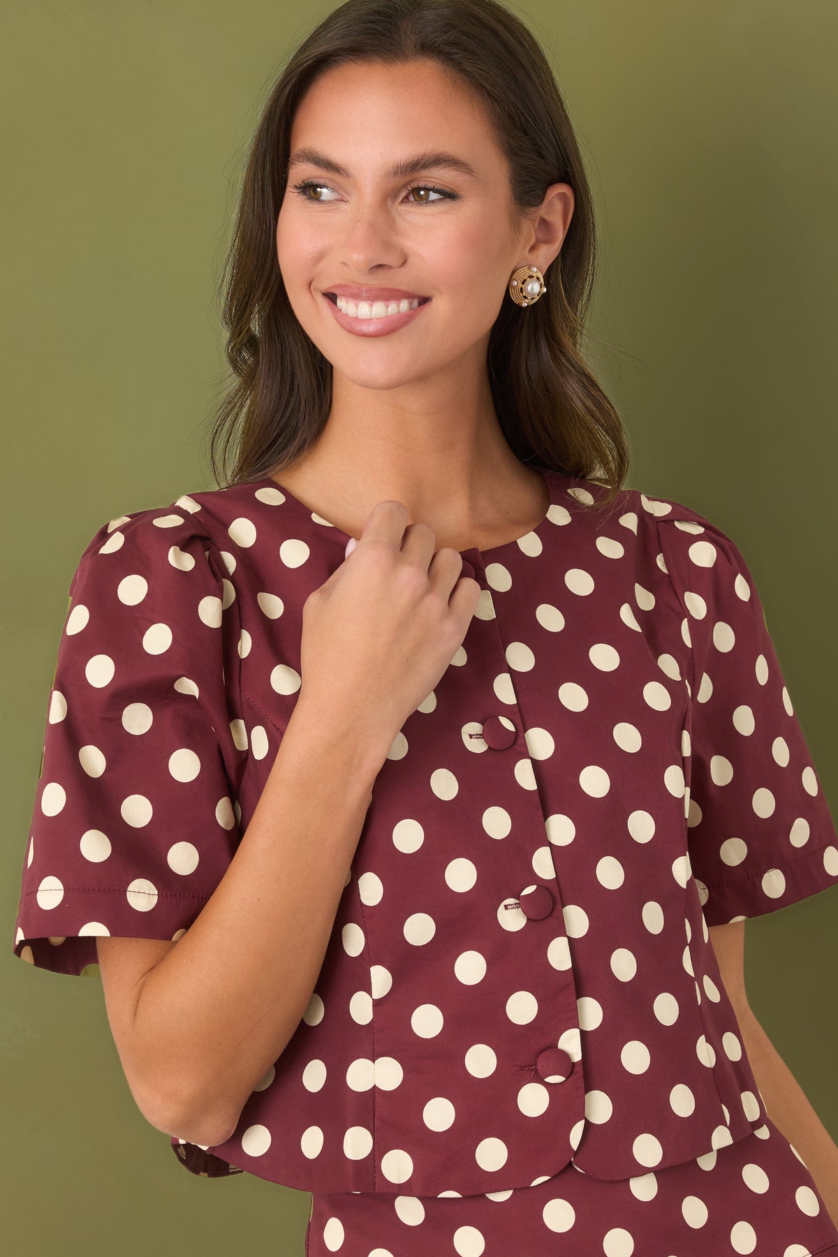 Crop in photo of a woman modeling a cropped, short sleeve, button up blouse. The blouse is a wine red color with ivory polka dots and she is standing against an olive backdrop.