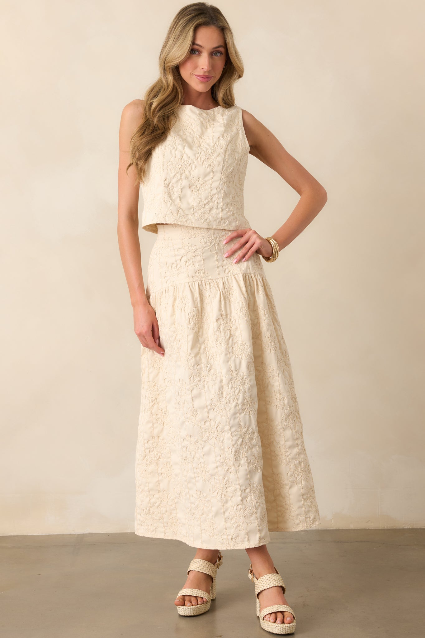 A full-body view of the beige sleeveless top, featuring a round neckline, back keyhole button closure, and monochrome floral embroidery throughout. The top has a slightly cropped length and a hidden side zipper.