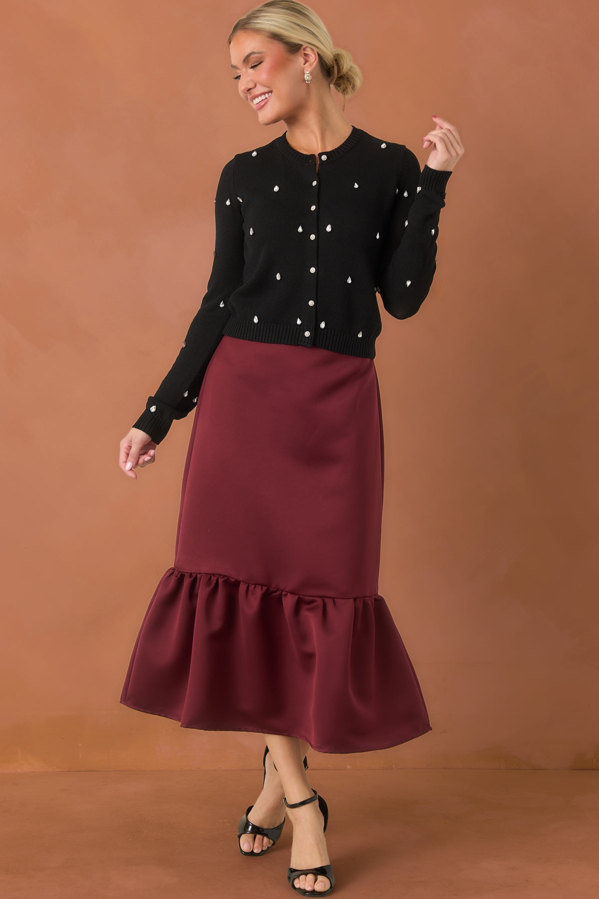 Woman modeling a rich burgundy midi skirt that has a loose, mermaid structure. It is paired with a black, rhinestone cardigan and she is standing in front of a burnt orange back drop.
