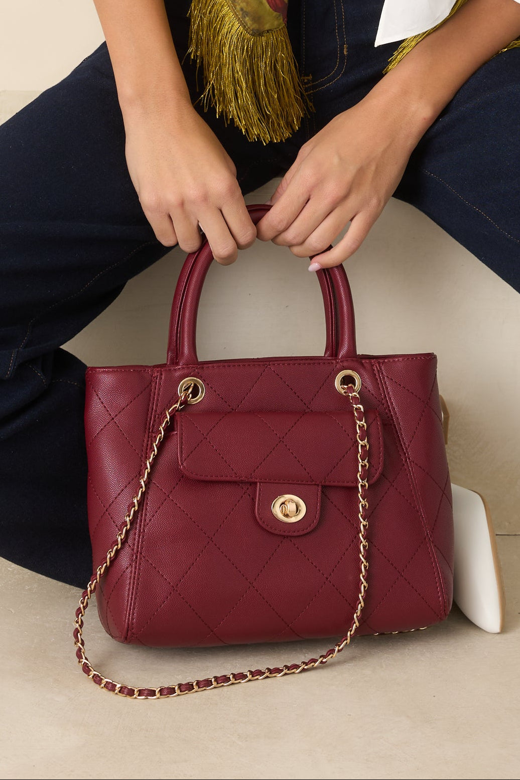 A burgundy quilted leather handbag with gold-tone hardware and a chain strap, held by a model wearing a white blouse, dark blue jeans, and a colorful scarf.