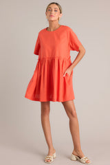 This orange dress features a round neckline, a keyhole cutout at the back of the neck with a button closure, short sleeves, ruffle detailing along the waistline, and functional pockets at the hips.