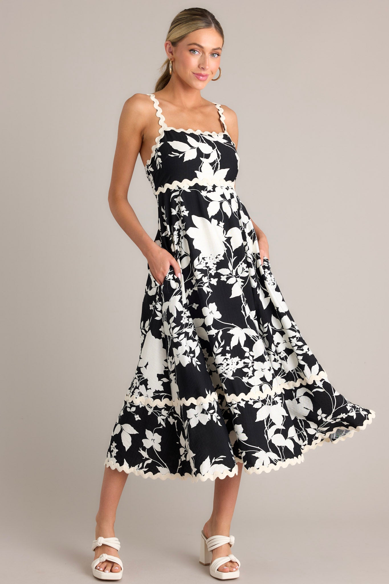 Angled full body view of this black floral midi dress that features a square neckline, thin ricrac straps, a fully smocked back, ricrac detailing, functional hip pockets, and a ricrac hemline.