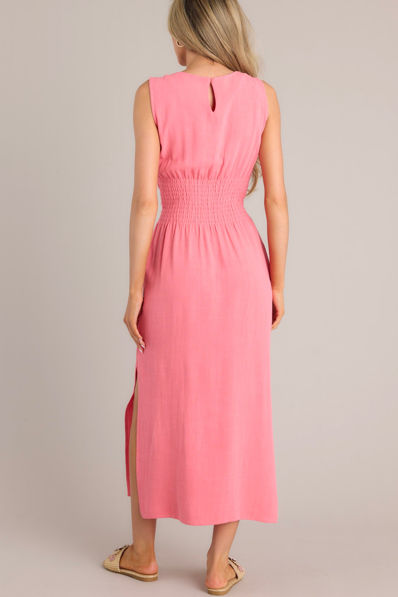 Back view of this coral dress that features a rounded neckline, a back keyhole cutout with functional button, a smocked back insert, a self-tie waist feature, functional hip pockets, and side slits.