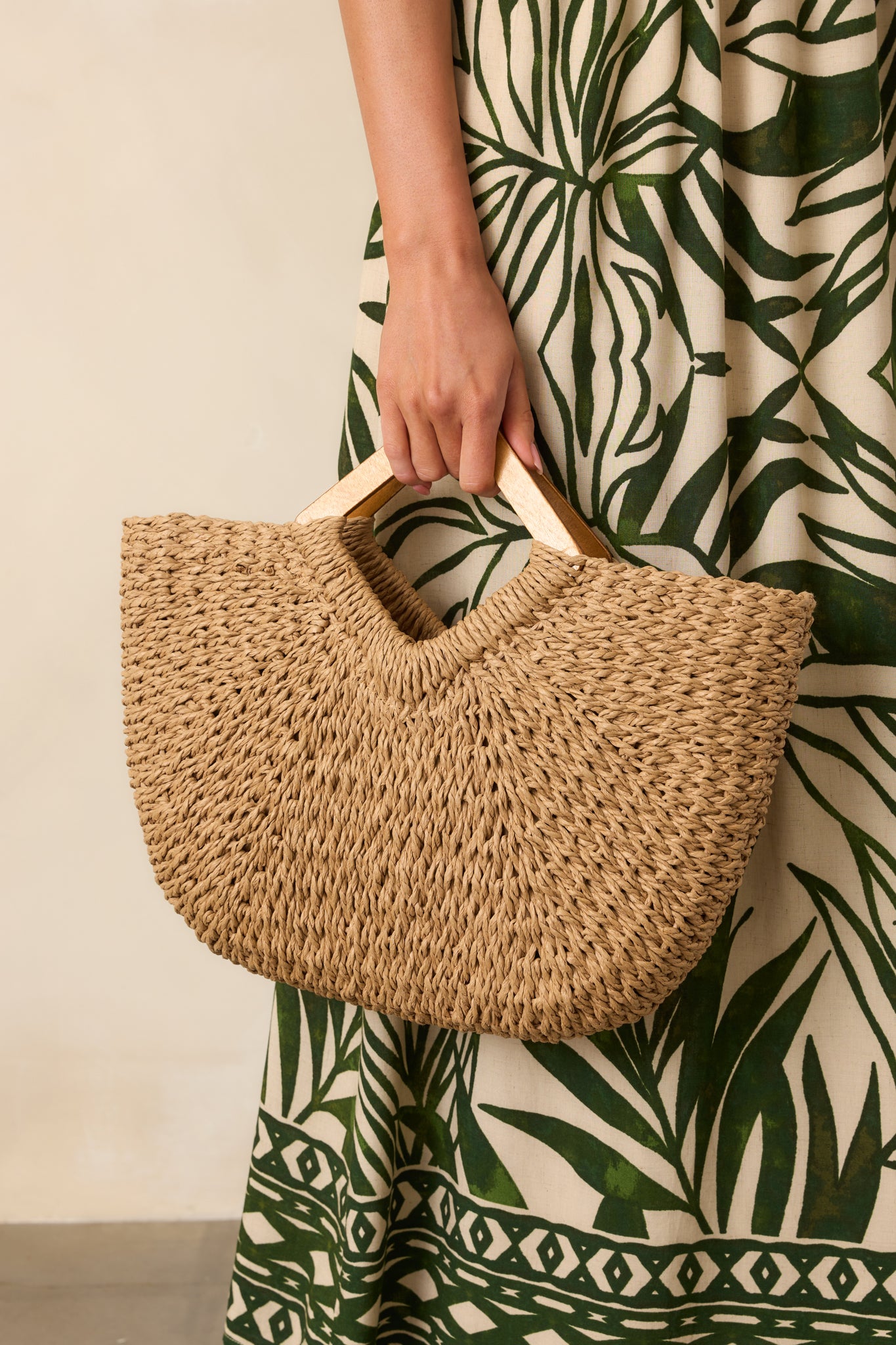 Straw-textured tan handbag with wooden handles displayed at full length, emphasizing its shape and natural color contrast with the outfit.