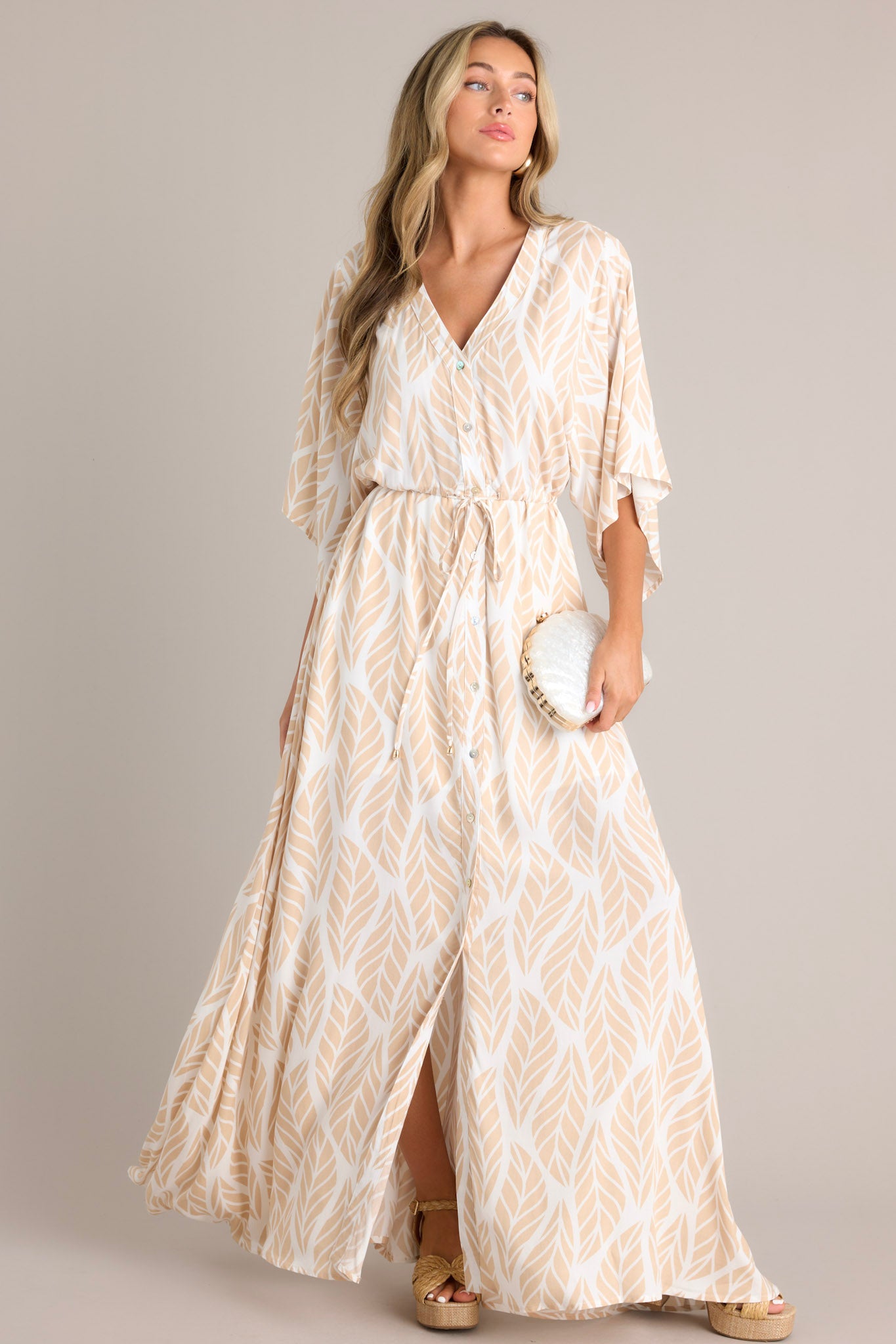 Angled full body view of this beige maxi dress that features a v-neckline, a functional button front, a self-tie drawstring waist, a flowing silhouette, and flowing half sleeves.