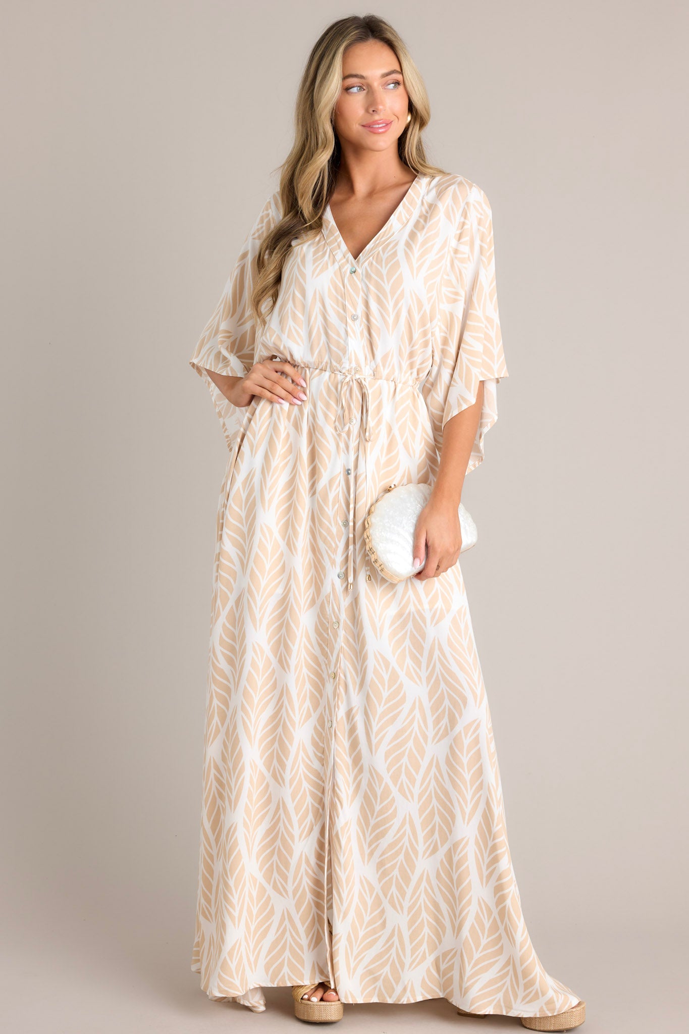 Full body view of this beige maxi dress that features a v-neckline, a functional button front, a self-tie drawstring waist, a flowing silhouette, and flowing half sleeves.