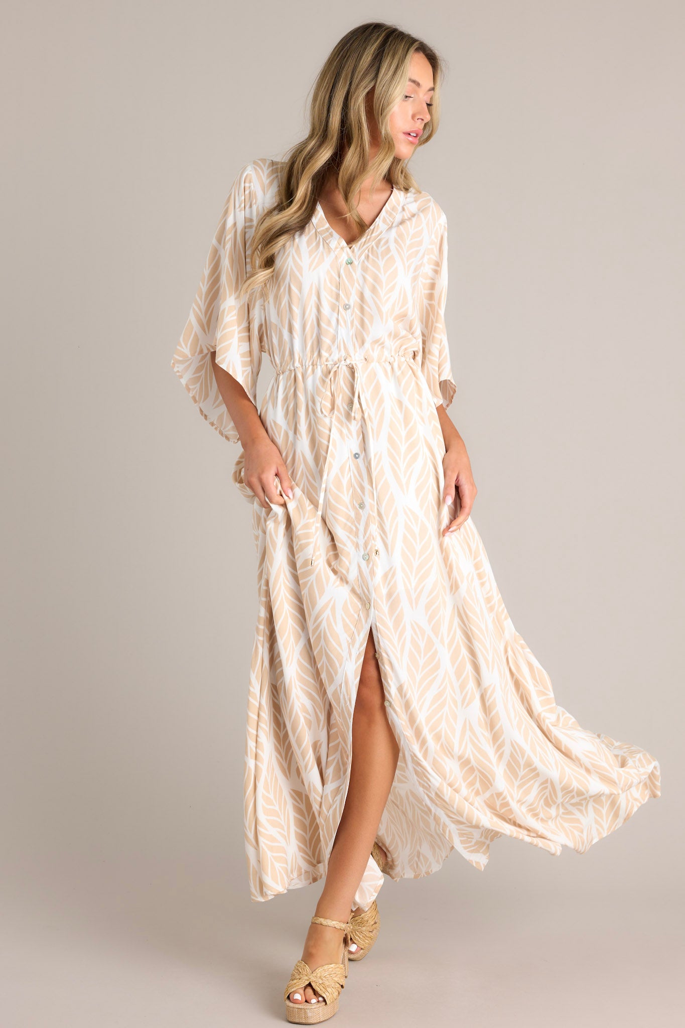 Front view of this beige maxi dress that features a v-neckline, a functional button front, a self-tie drawstring waist, a flowing silhouette, and flowing half sleeves.