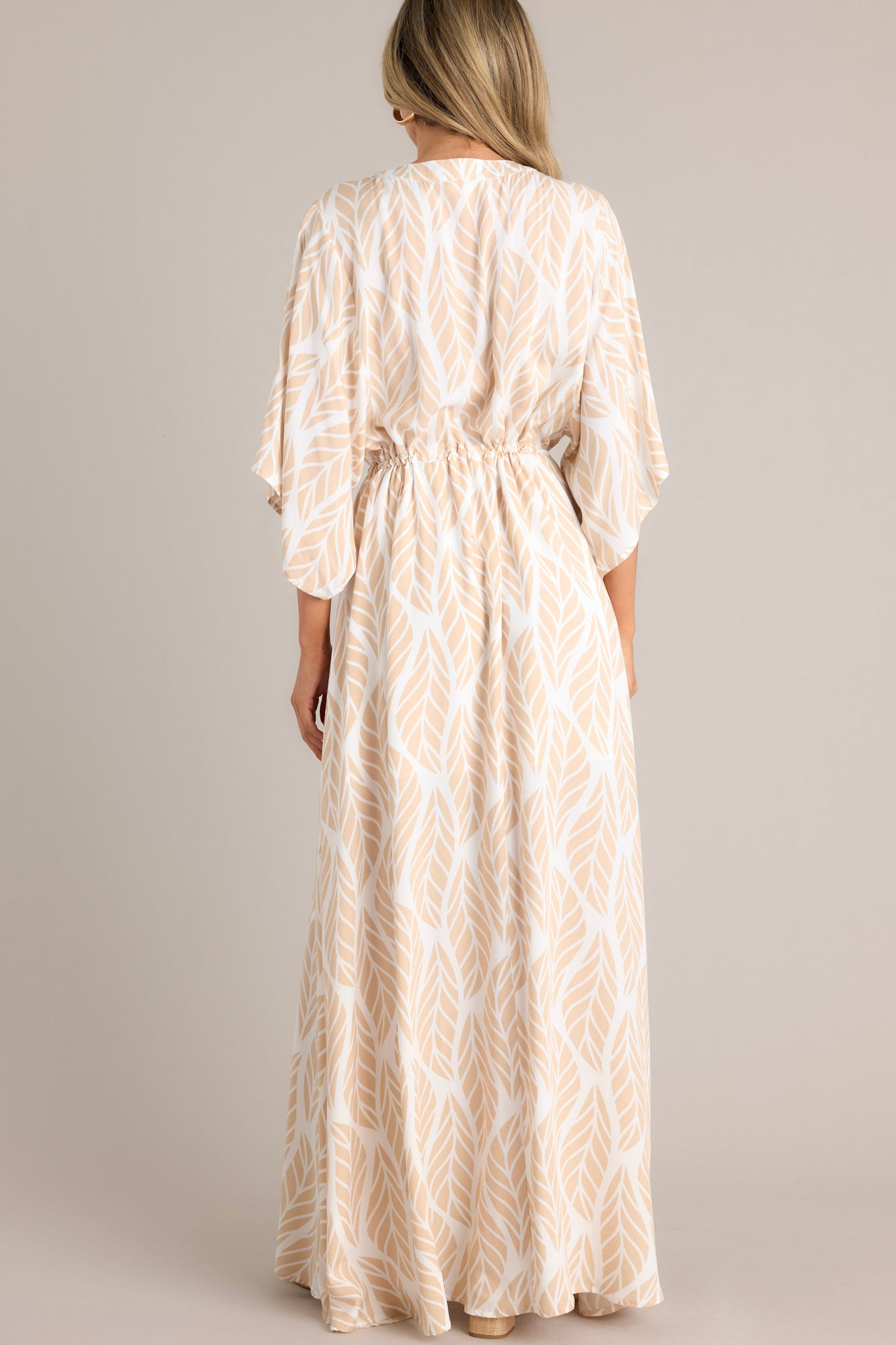 Back view of this beige maxi dress that features a v-neckline, a functional button front, a self-tie drawstring waist, a flowing silhouette, and flowing half sleeves.