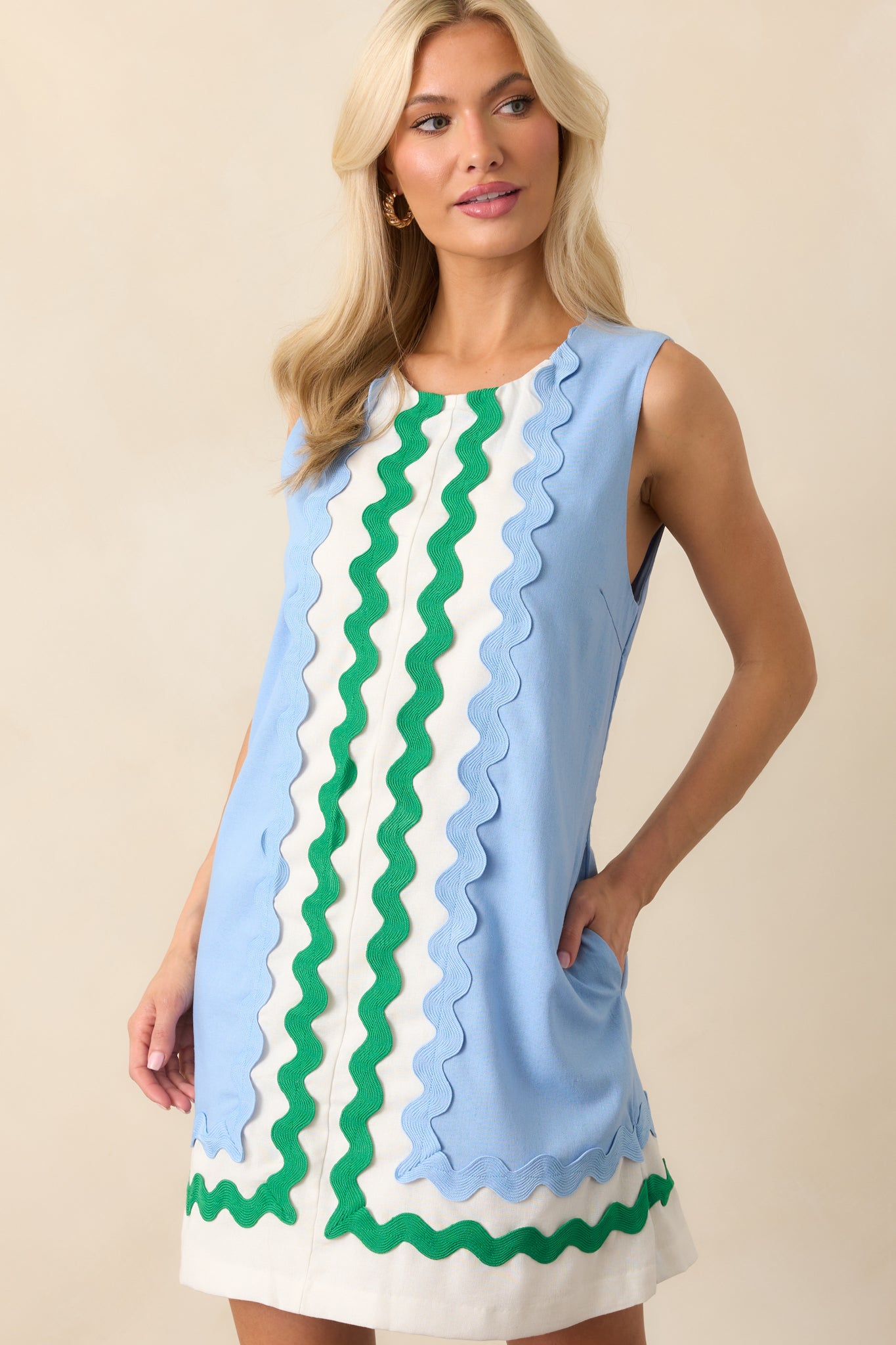 Up-close photo of model wearing light blue mini dress features white contrast panels at the center and hem, green and blue ric rac trim, a sleeveless design, round neckline, hidden back zipper, shift silhouette, and functional pockets.