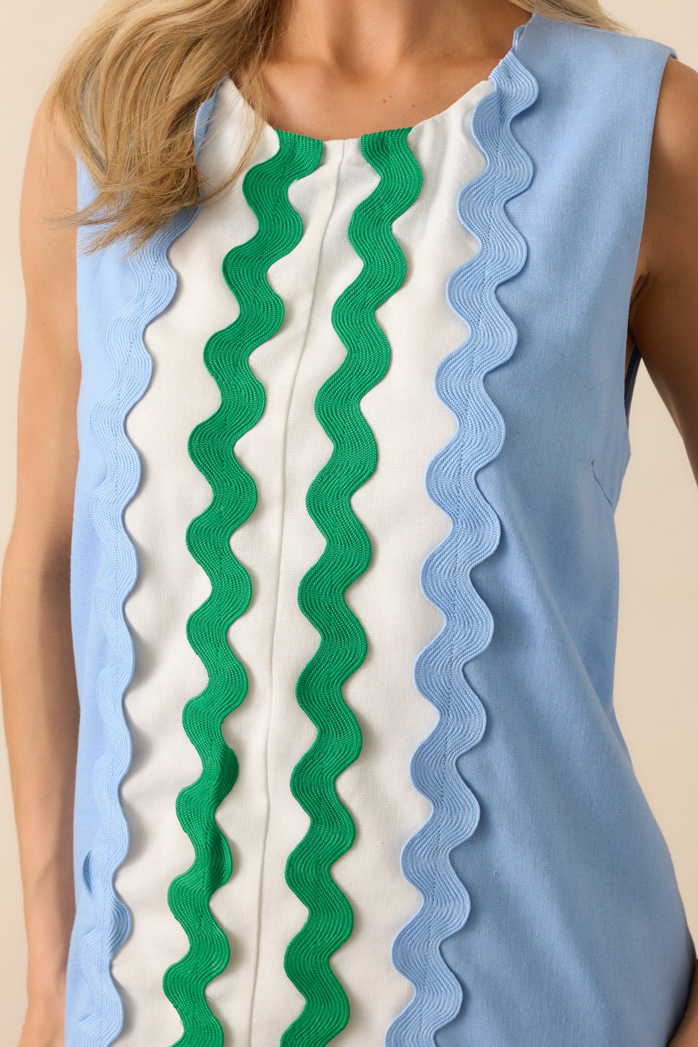 Up-close photo of model wearing light blue mini dress features white contrast panels at the center and hem, green and blue ric rac trim, a sleeveless design, round neckline, hidden back zipper, shift silhouette, and functional pockets.