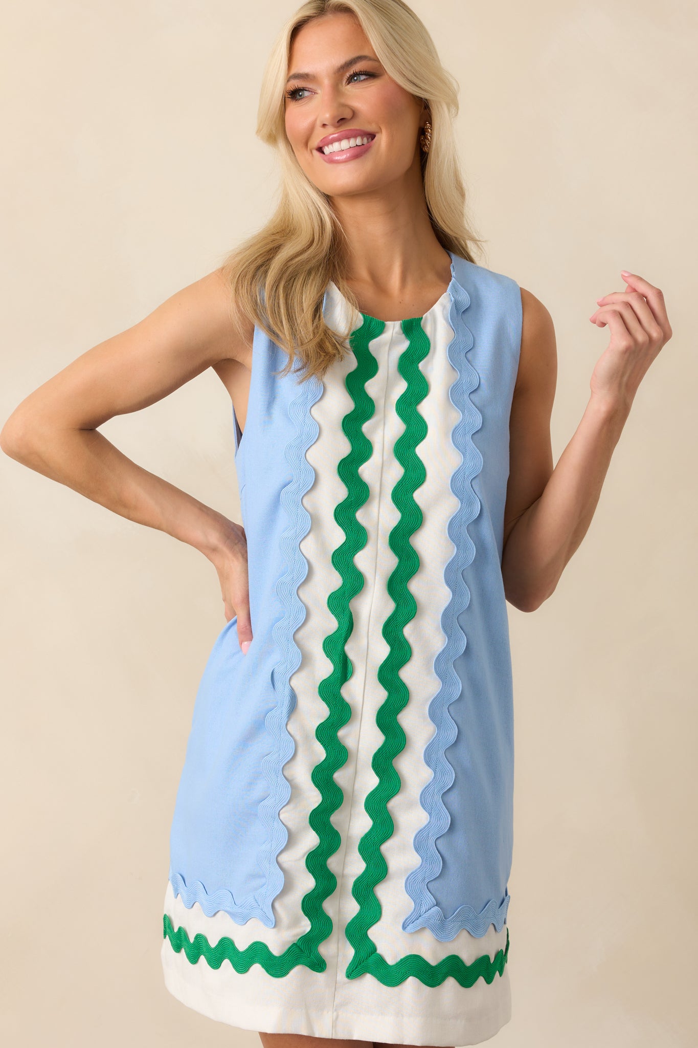 Up-close photo of model wearing light blue mini dress features white contrast panels at the center and hem, green and blue ric rac trim, a sleeveless design, round neckline, hidden back zipper, shift silhouette, and functional pockets.
