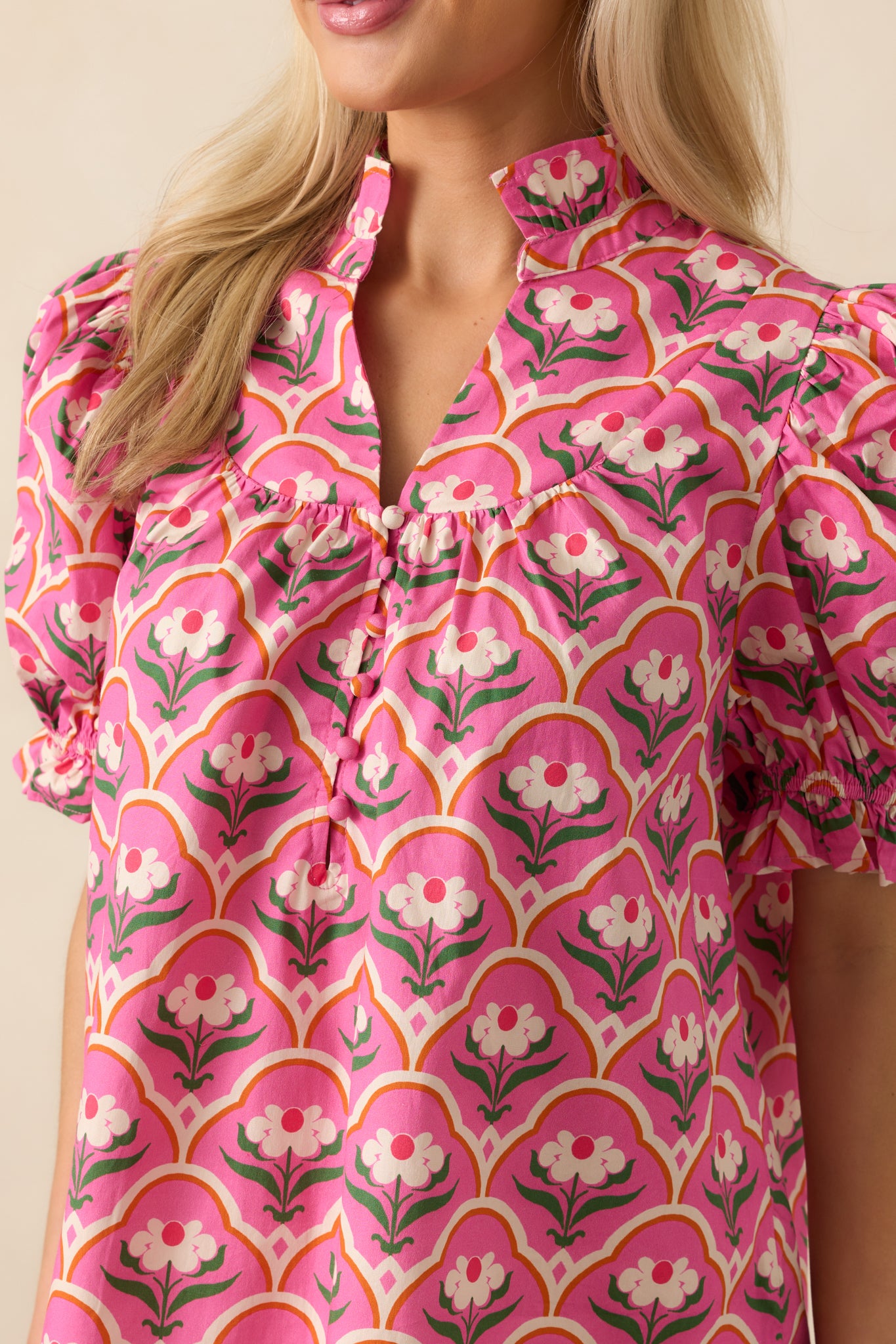 Focused crop of pink top that features a pink multicolor geometric floral print, ruffle notch v-neckline, functional button front, and short puff sleeves.